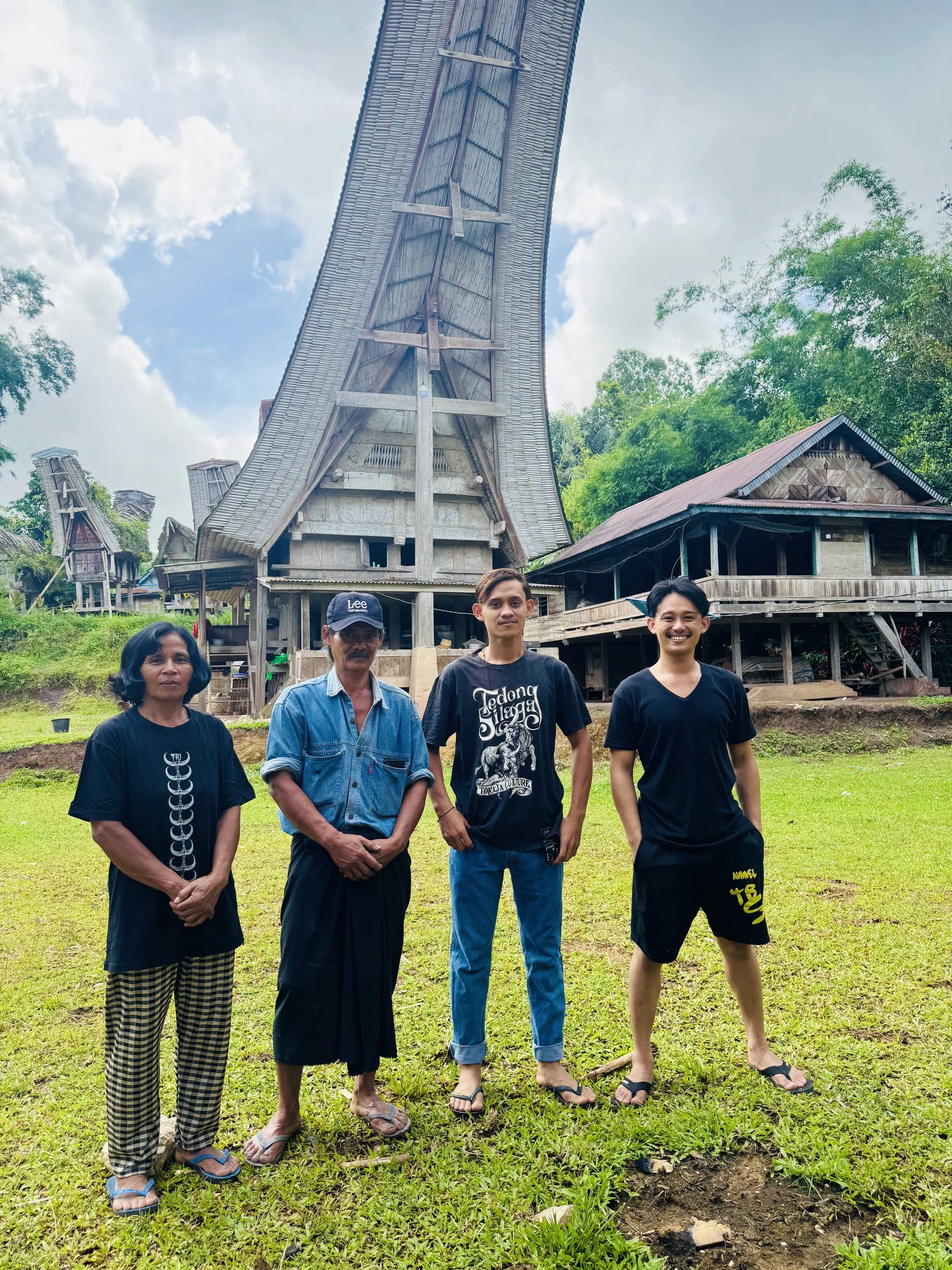 Four people standing on the grass in front of traditional wooden houses with tall, curved roofs, surrounded by green trees under a partly cloudy sky.
