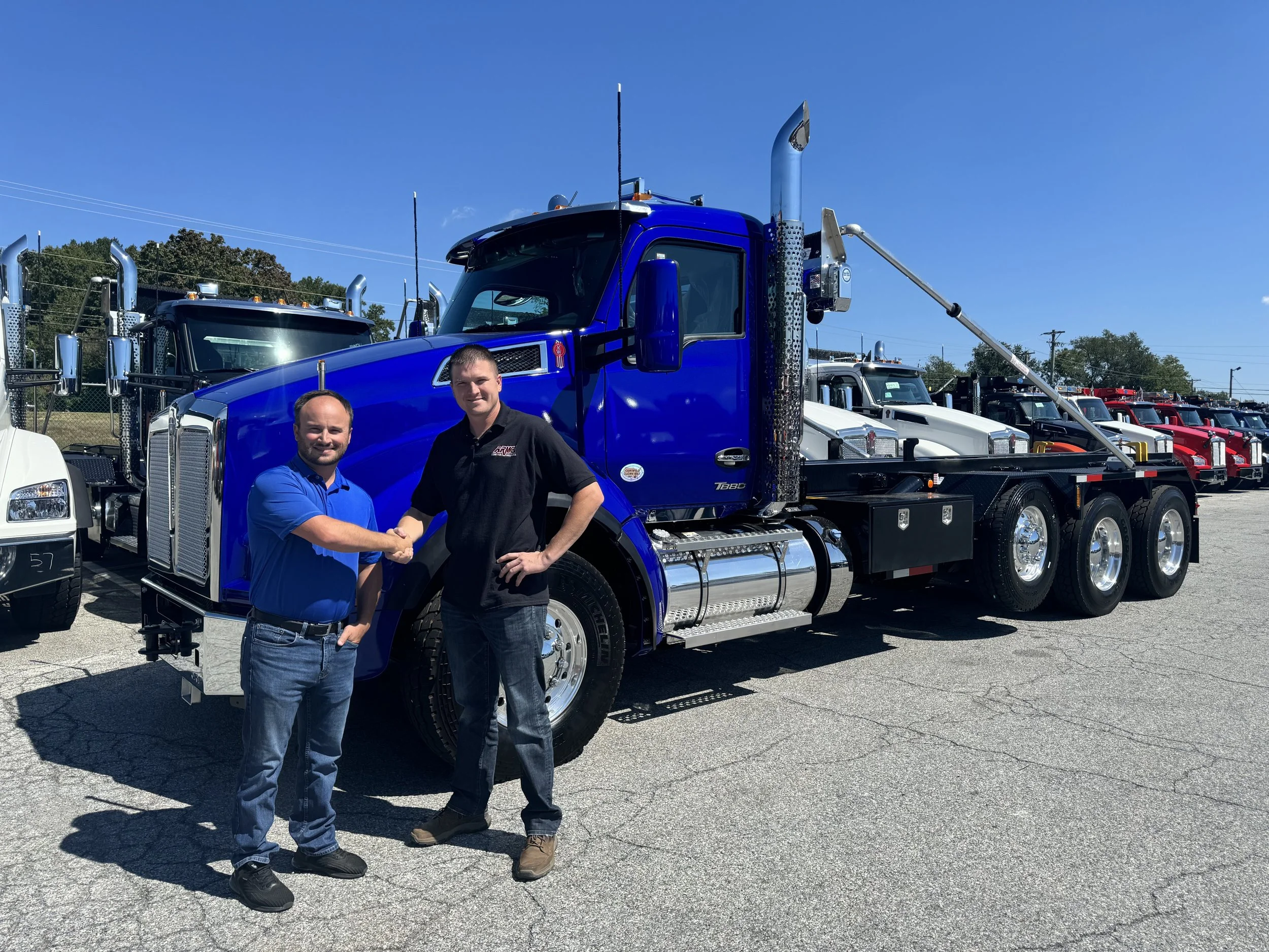 Armco Scrap Metals LLC owner Chris Lear shaking hands in front of a large blue tow truck with a flatbed, parked in a lot with multiple trucks.
