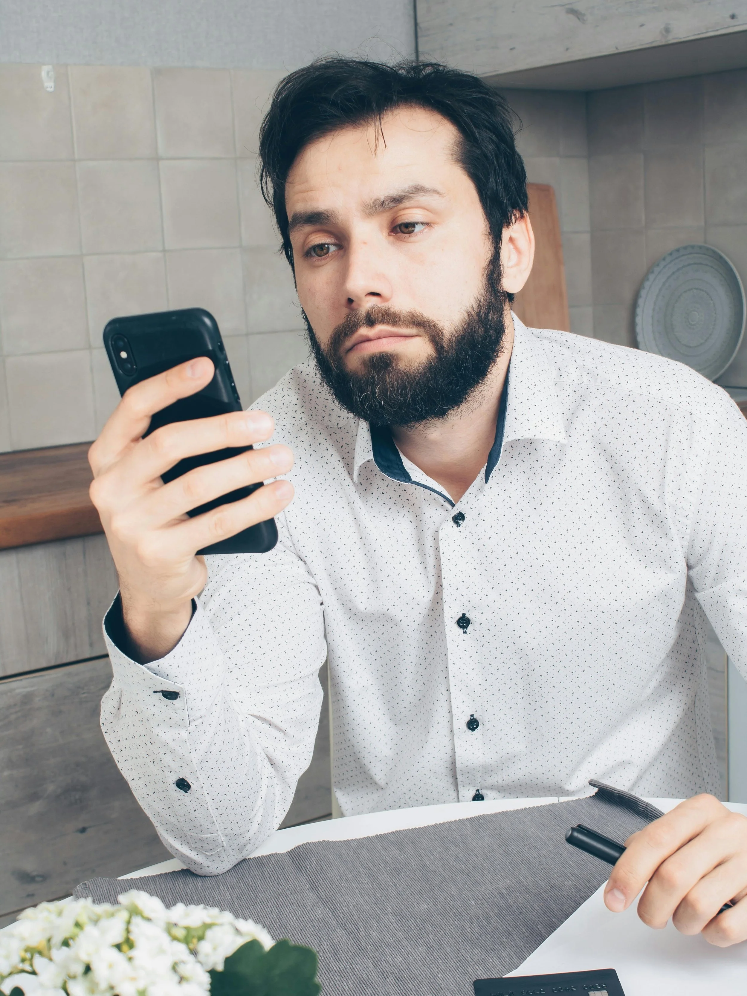 A man with dark hair and a beard sitting at a table, looking at a smartphone in his hand, appearing frustrated because he can't find a mobile notary public in Indianapolis