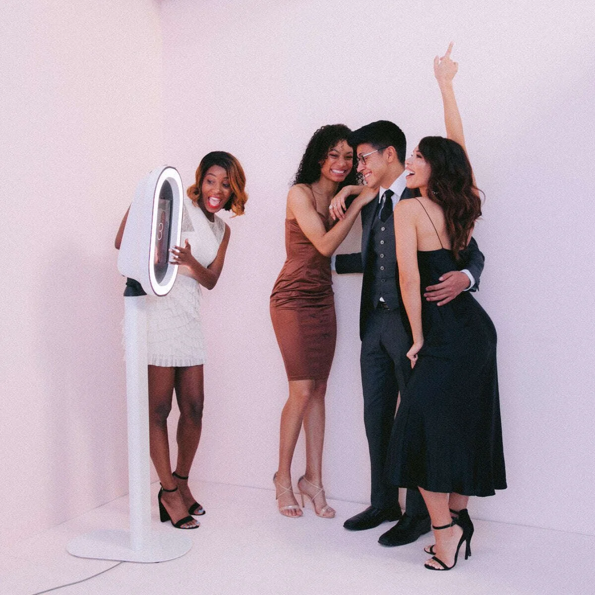Four friends in fancy clothes taking photos and enjoying each other's company against a plain pink wall.