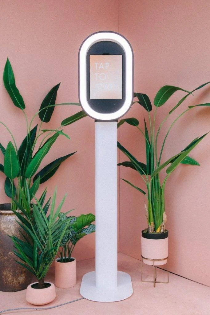 A tall, white, digital kiosk with a rounded rectangular screen displaying 'TAP TO' in white text, standing on a pink floor against a pink wall. The kiosk is surrounded by green houseplants in pink and beige pots.
