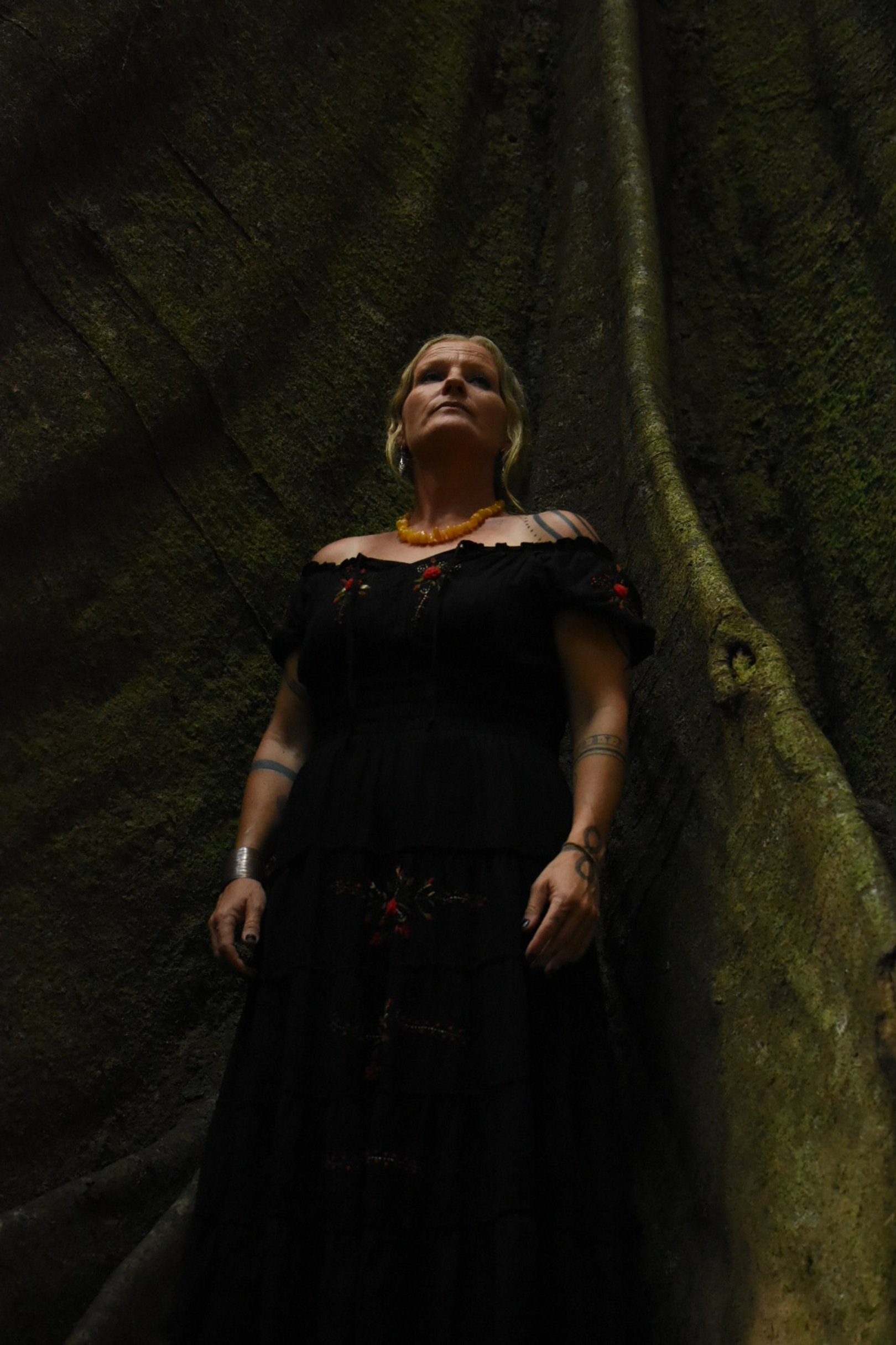 A woman standing in a dark, moss-covered cave or tunnel, looking upwards with a serious expression, wearing a black off-shoulder dress, orange necklace, and various tattoos on her arms.