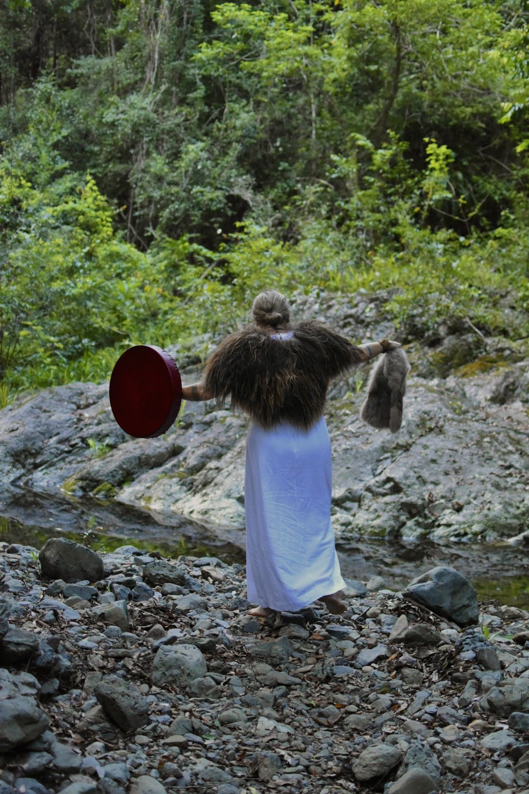 A woman dressed in white with a fur shawl standing on rocky ground near a stream, holding a red shield in her left hand and a fur pouch in her right hand, facing away from the camera in a green forest.