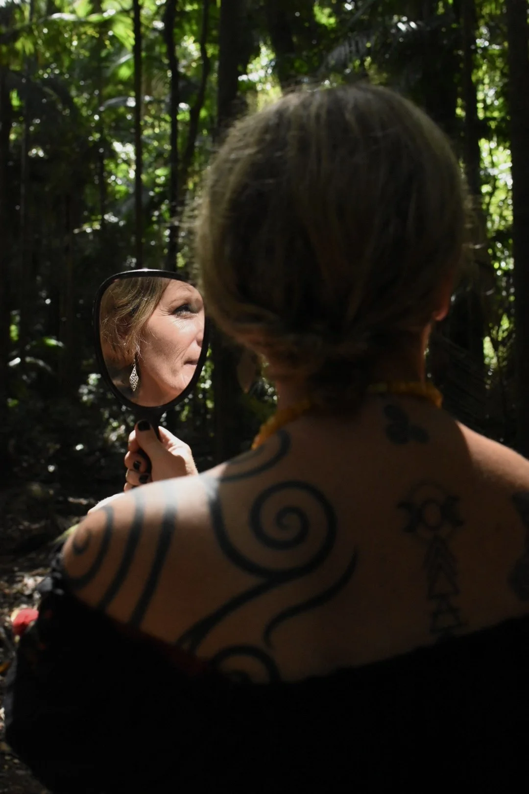 A woman with tattoos on her shoulder and back holding a mirror while standing in a forest, reflecting her face.