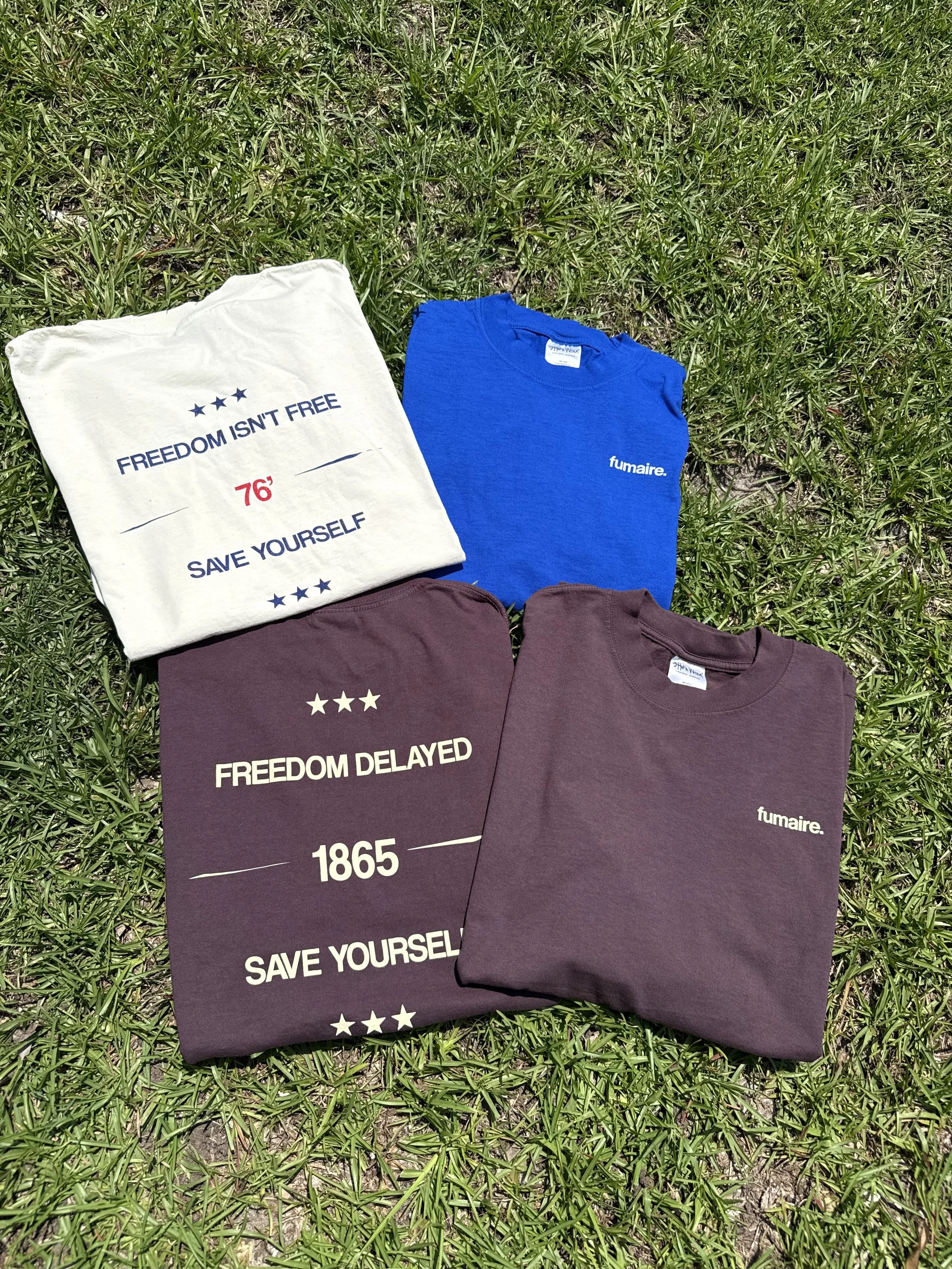 A collection of folded t-shirts lying on grass. One white shirt with text reading 'FREEDOM ISN'T FREE', '76', and 'SAVE YOURSELF'. Two maroon shirts with text reading 'FREEDOM DELAYED' and '1865'. A blue shirt with a small white logo reading 'fumaire.'.