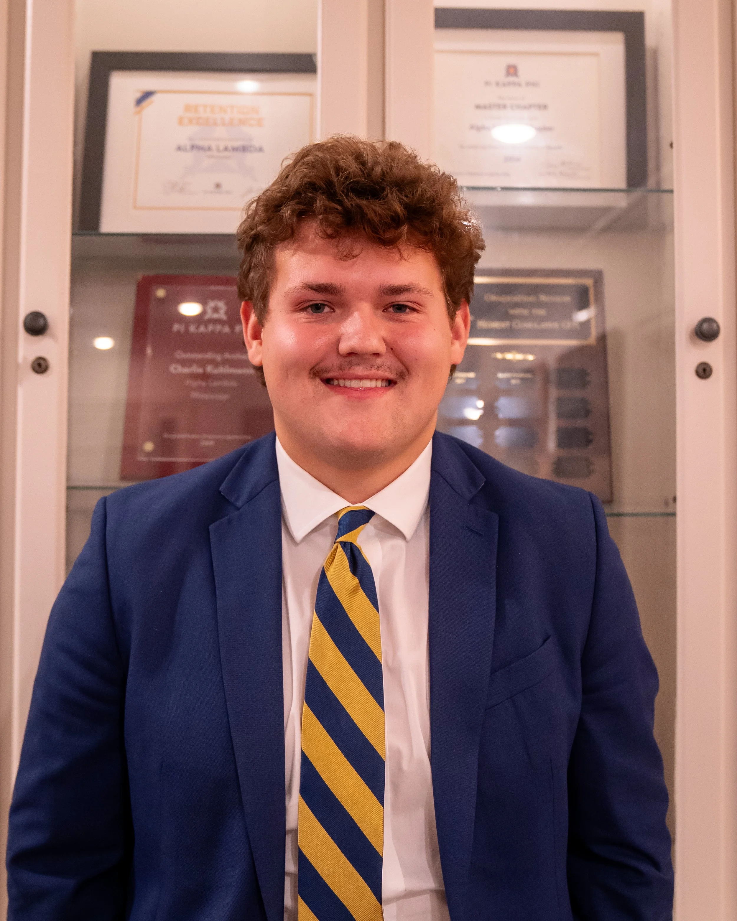 A young man wearing a black suit with a white shirt and red tie, standing in front of a flag and wooden stairs, smiling at the camera.