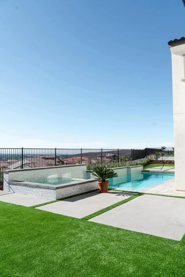 Backyard with swimming pool, spa, and artificial grass, fence in background, clear sky.