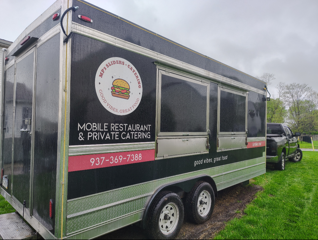 Black food truck with a sign advertising mobile restaurant and private catering, featuring a hamburger logo and the phrase 'good vibes, great food', parked on grass with a black pickup truck nearby and cloudy sky above.