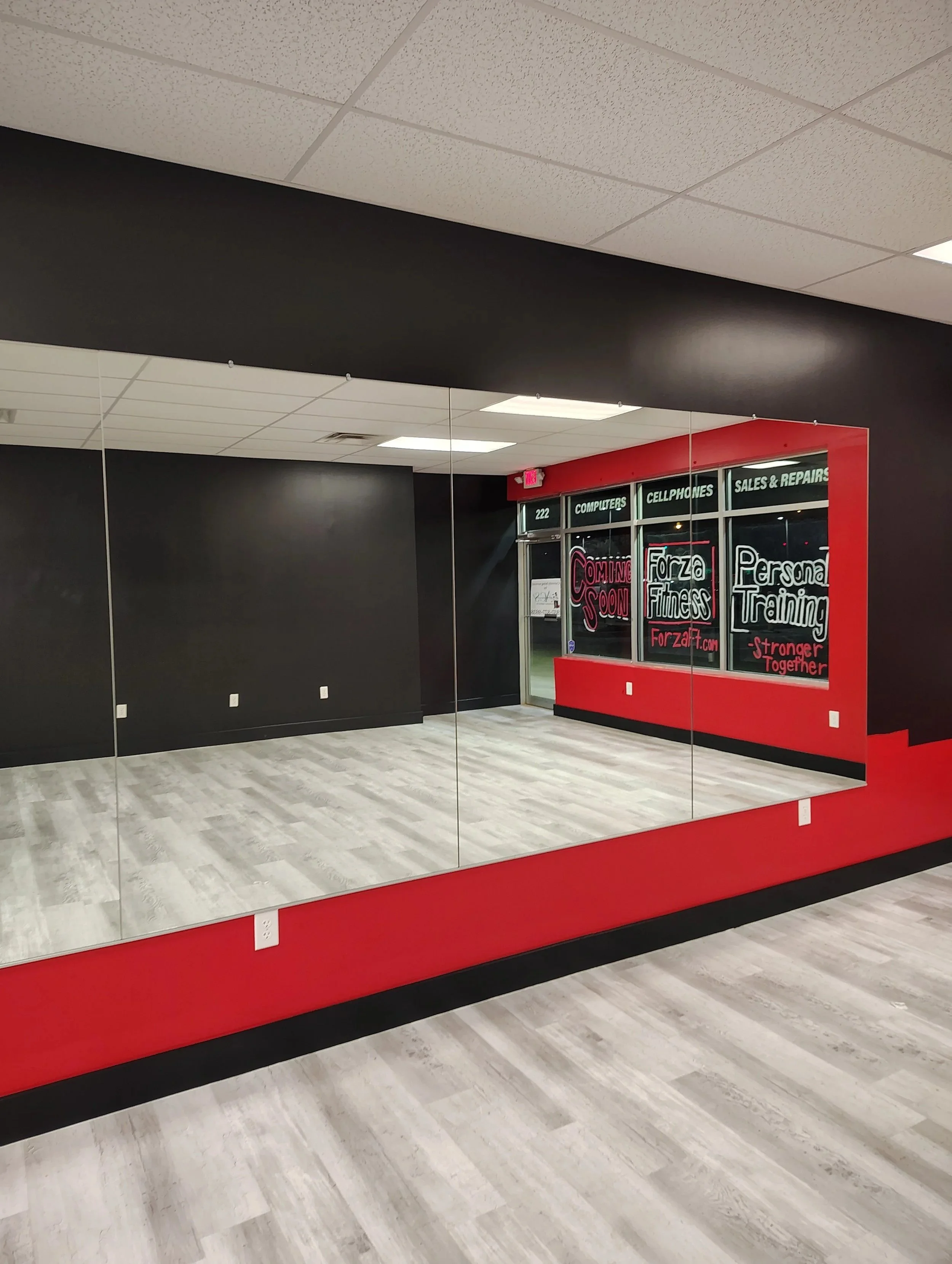 Empty indoor space with a large mirror on the wall, a red and black frame around a window advertising fitness and training services, and light-colored wood flooring.