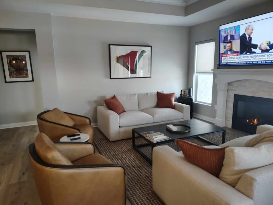Living room with a white sofa, two tan armchairs, a black coffee table, a fireplace, and a wall-mounted TV showing a news program.
