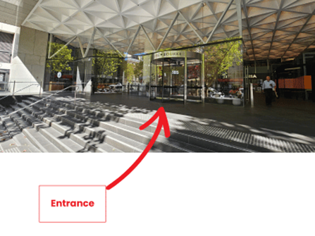 View of building entrance with glass doors, surrounded by open space and trees, under a geometric roof structure.