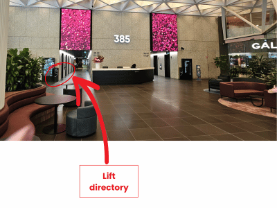 Interior of a building lobby with seating areas, plants, and digital screens on the walls. A red arrow points to a lift directory on the wall.