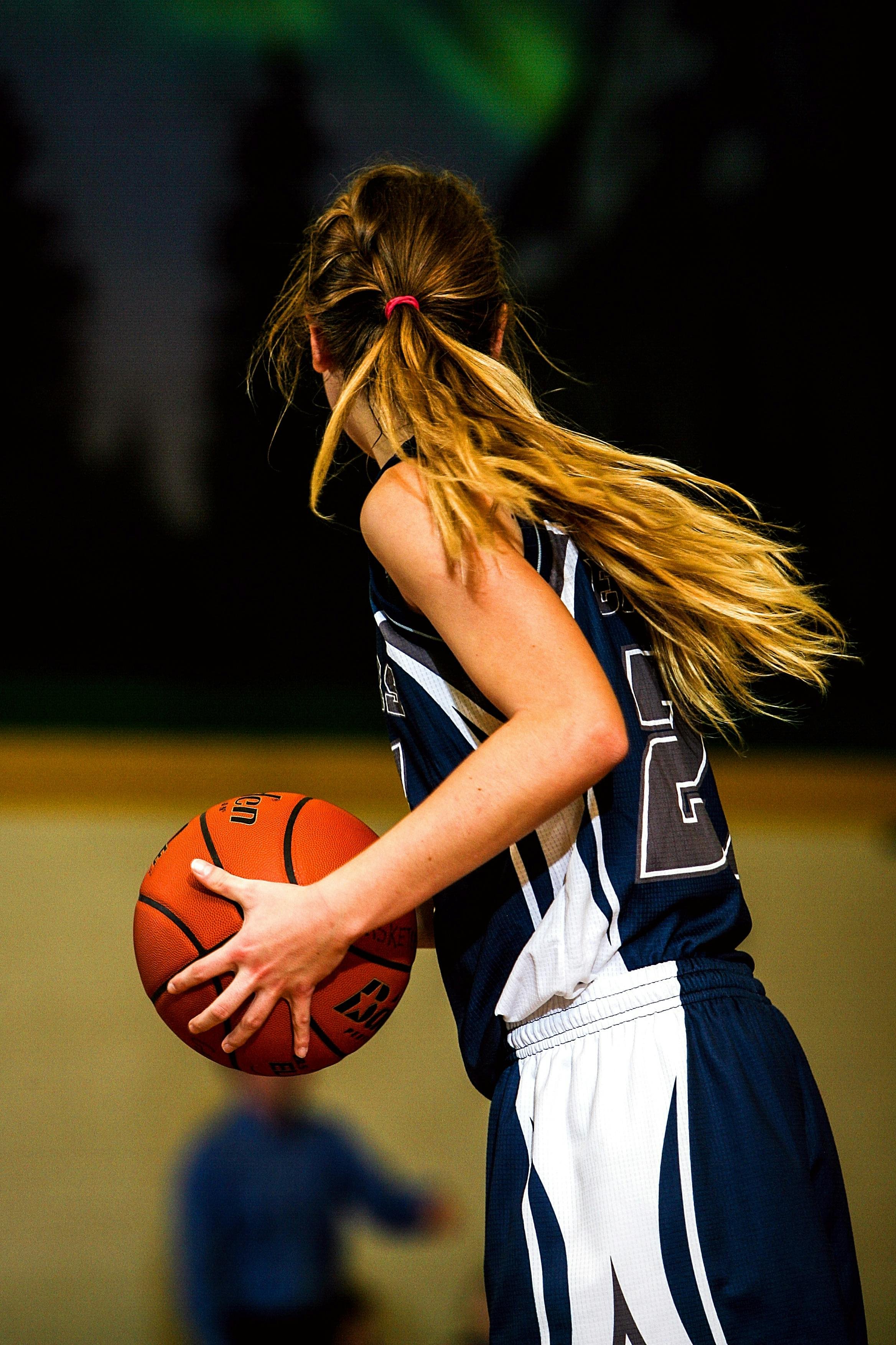 Young female basketball player in blue and white uniform holding a basketball, with long hair tied back in a ponytail, during an indoor game.