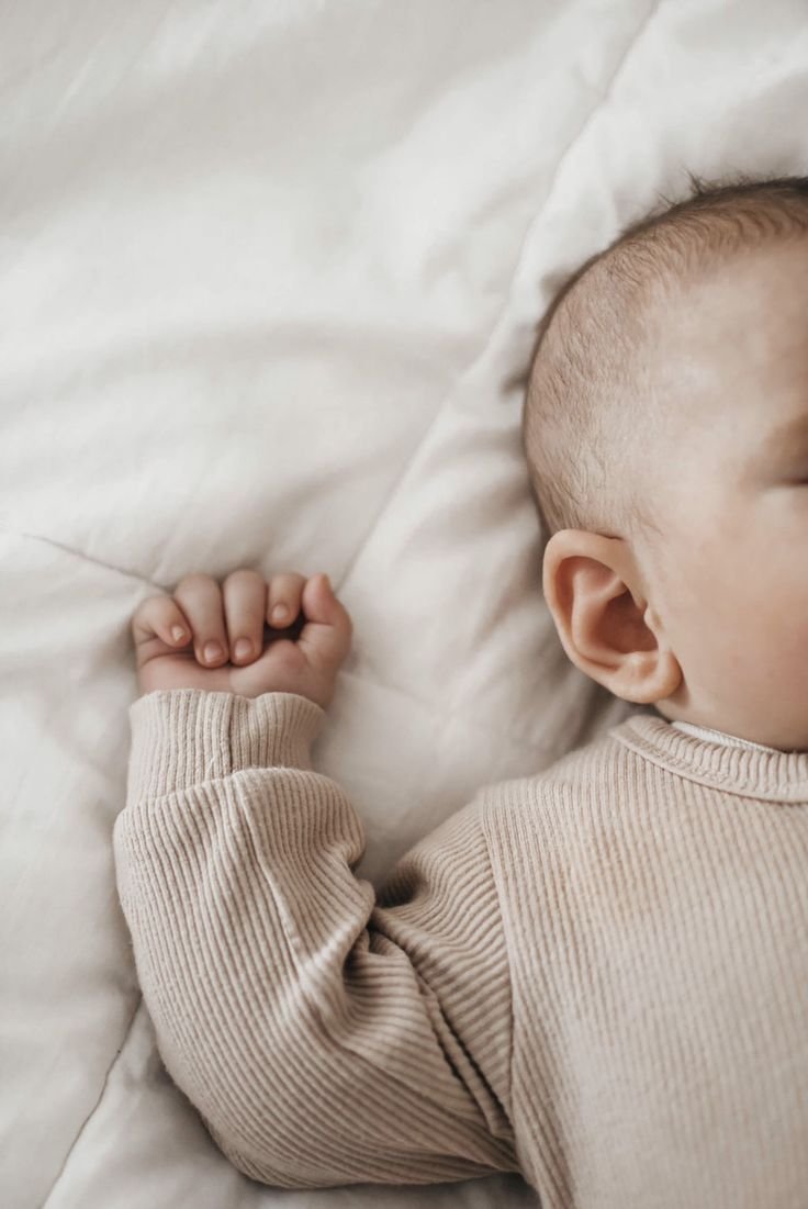 Close-up of a sleeping baby in a beige ribbed sweater, lying on a white blanket with one hand visible.