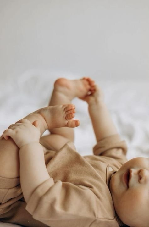 A baby in beige clothing lying on a soft surface, holding one foot with its hands.