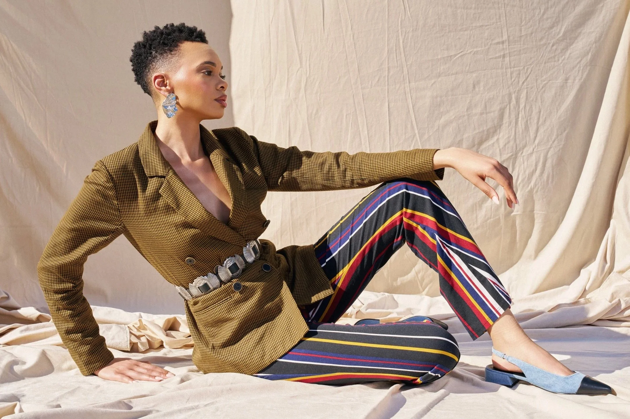 A woman sitting on the floor in front of a beige backdrop, wearing a brown blazer, colorful striped pants, and blue shoes. She is accessorized with large, shiny earrings and a decorative belt, with her hair styled short and natural.
