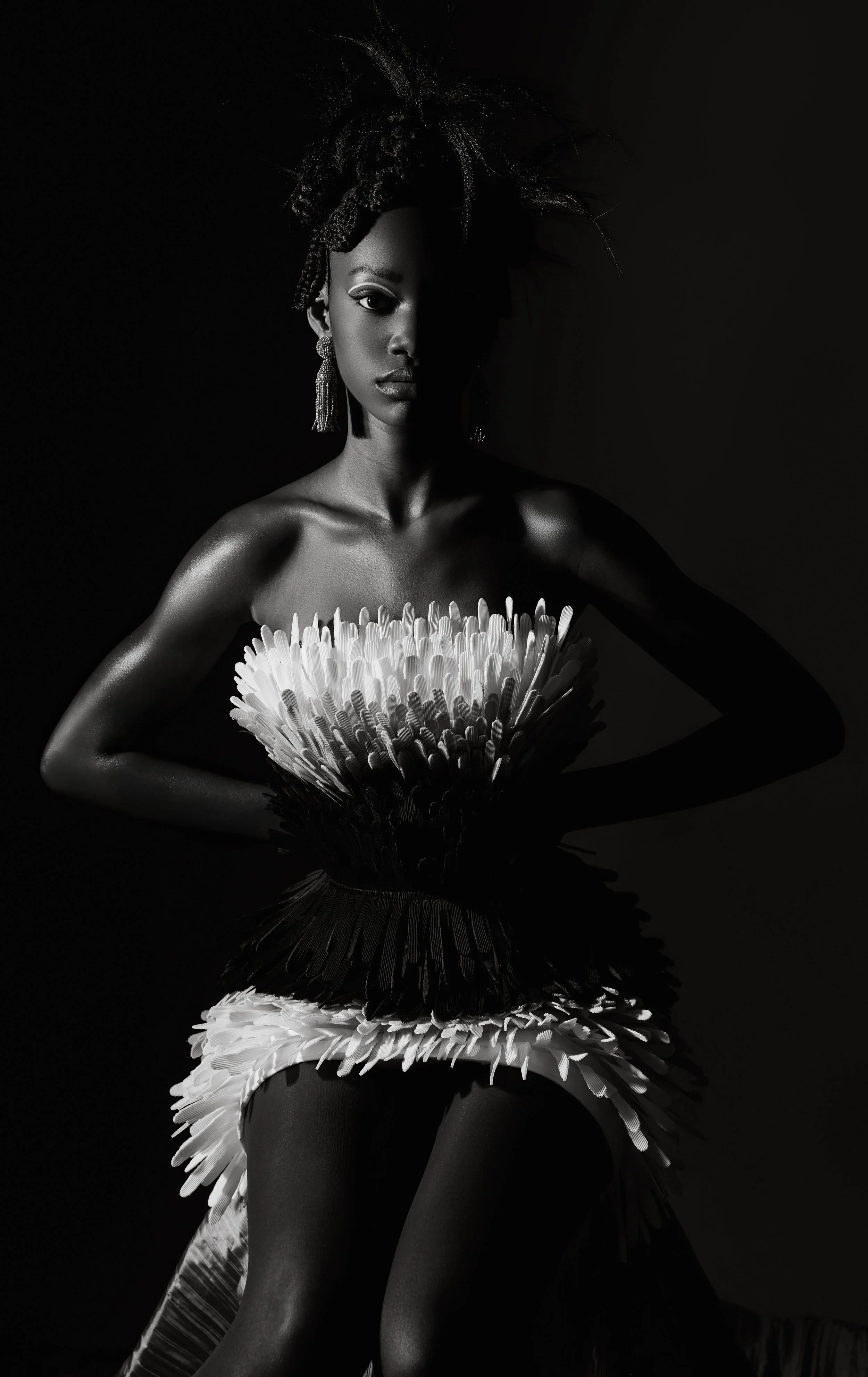 Black and white portrait of a woman with dramatic makeup and elaborate hairstyle, wearing statement earrings, and dressed in an artistic dress made of feather-like material, posing against a dark background with a reflective surface.