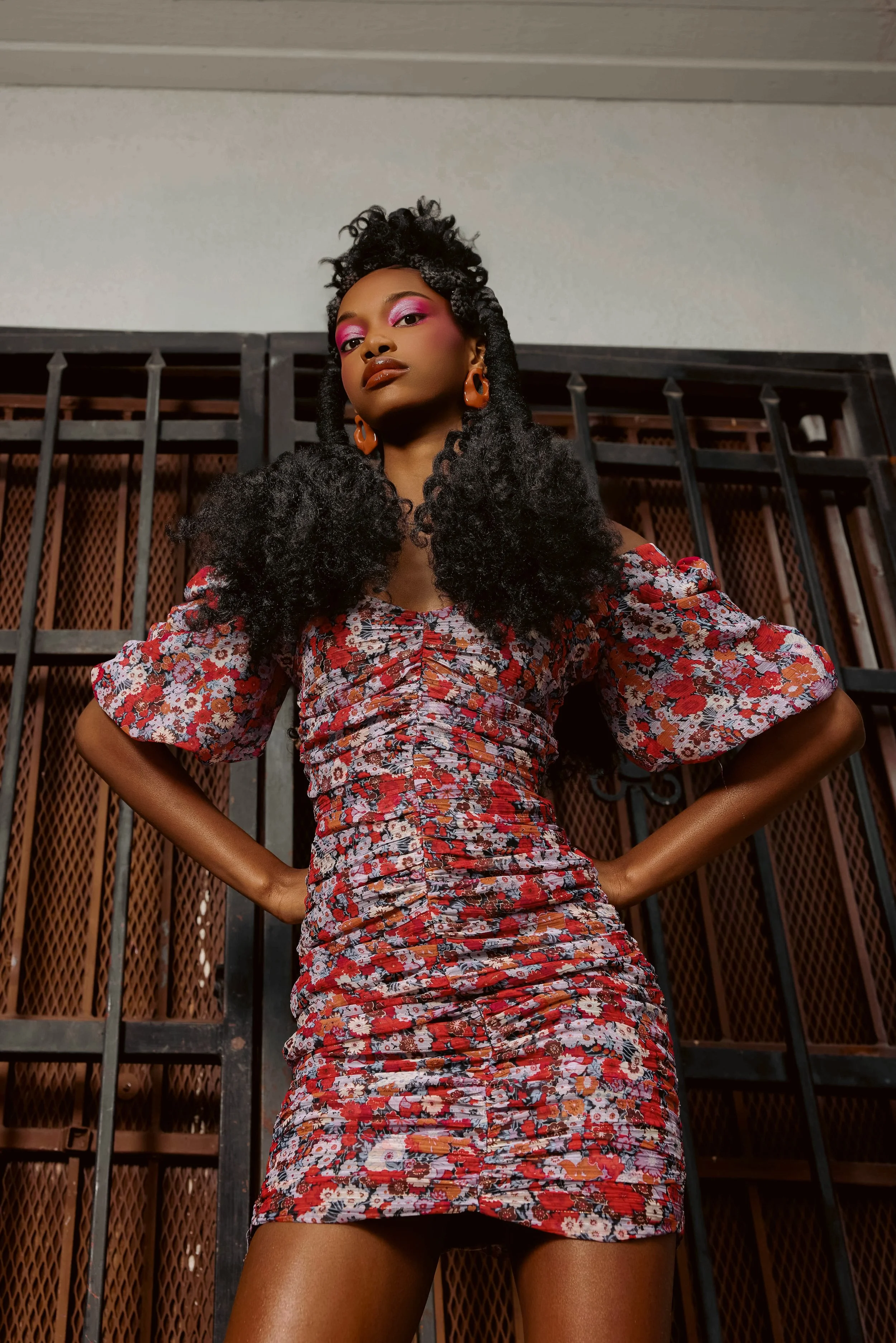 A woman with dark curly hair styled in an updo, wearing a floral dress with puffed sleeves, bold makeup featuring pink eyeshadow, and large orange earrings, standing confidently in front of a black iron gate.
