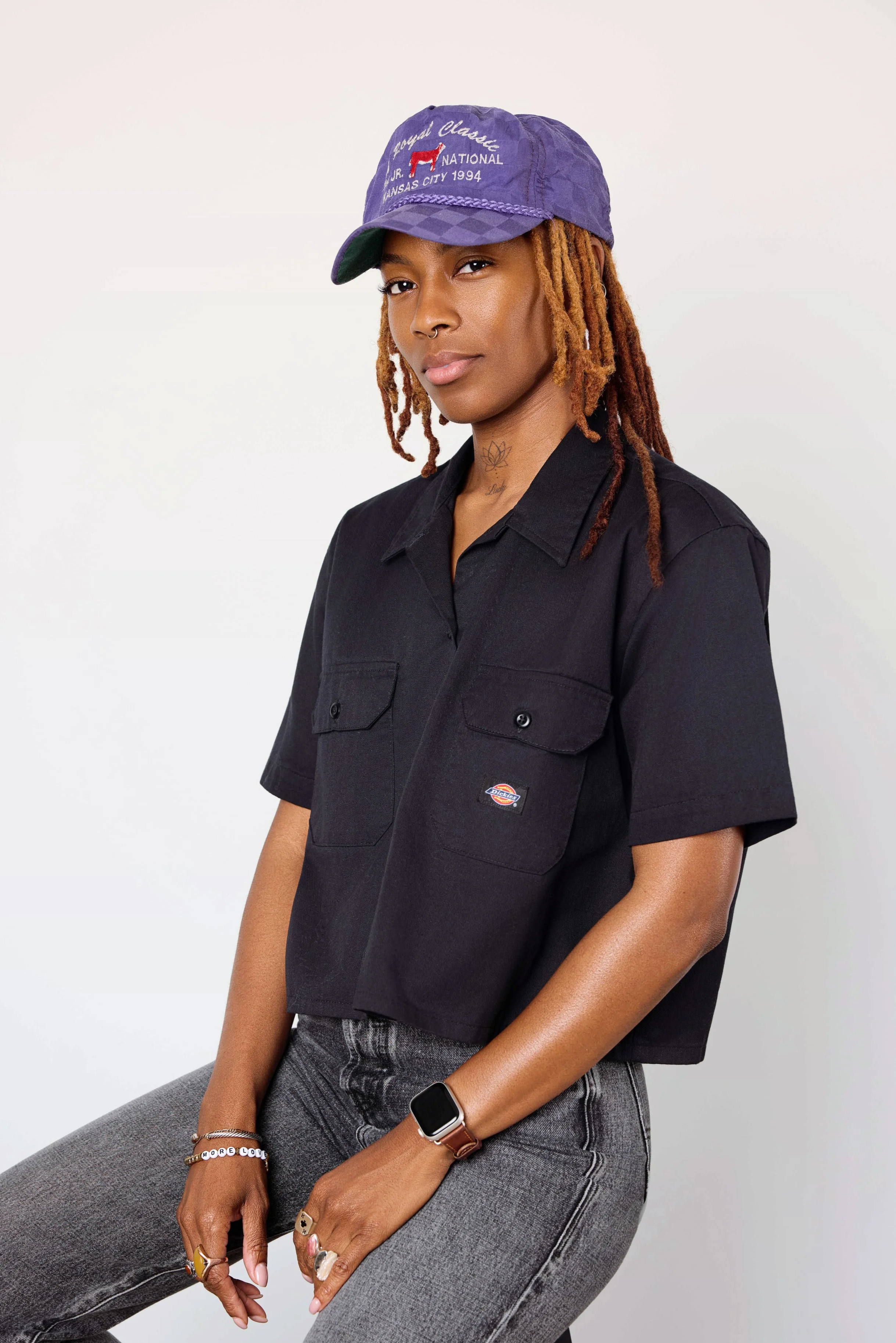 Portrait of a young woman with locs wearing a purple baseball cap, a black short-sleeve shirt, gray jeans, and various rings and bracelets, sitting against a white background.