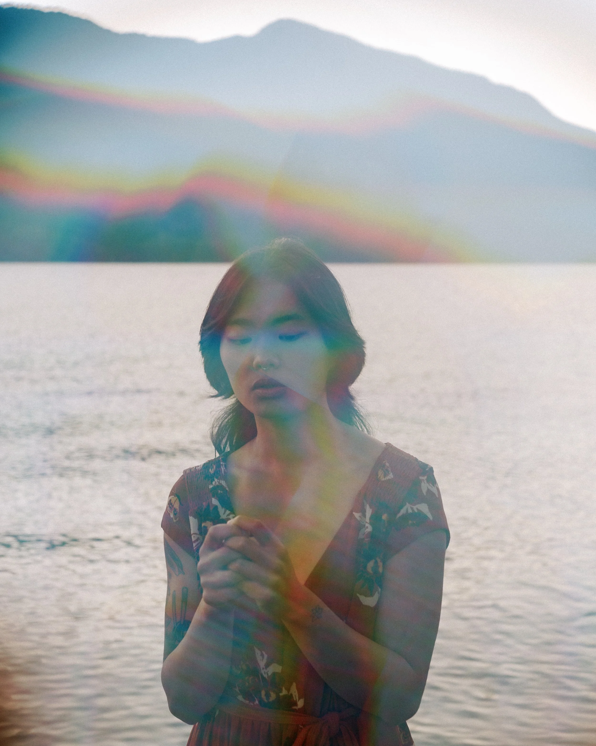 A woman standing by a body of water with mountains in the background, wearing a floral dress, with rainbow-colored lens flare across the image.