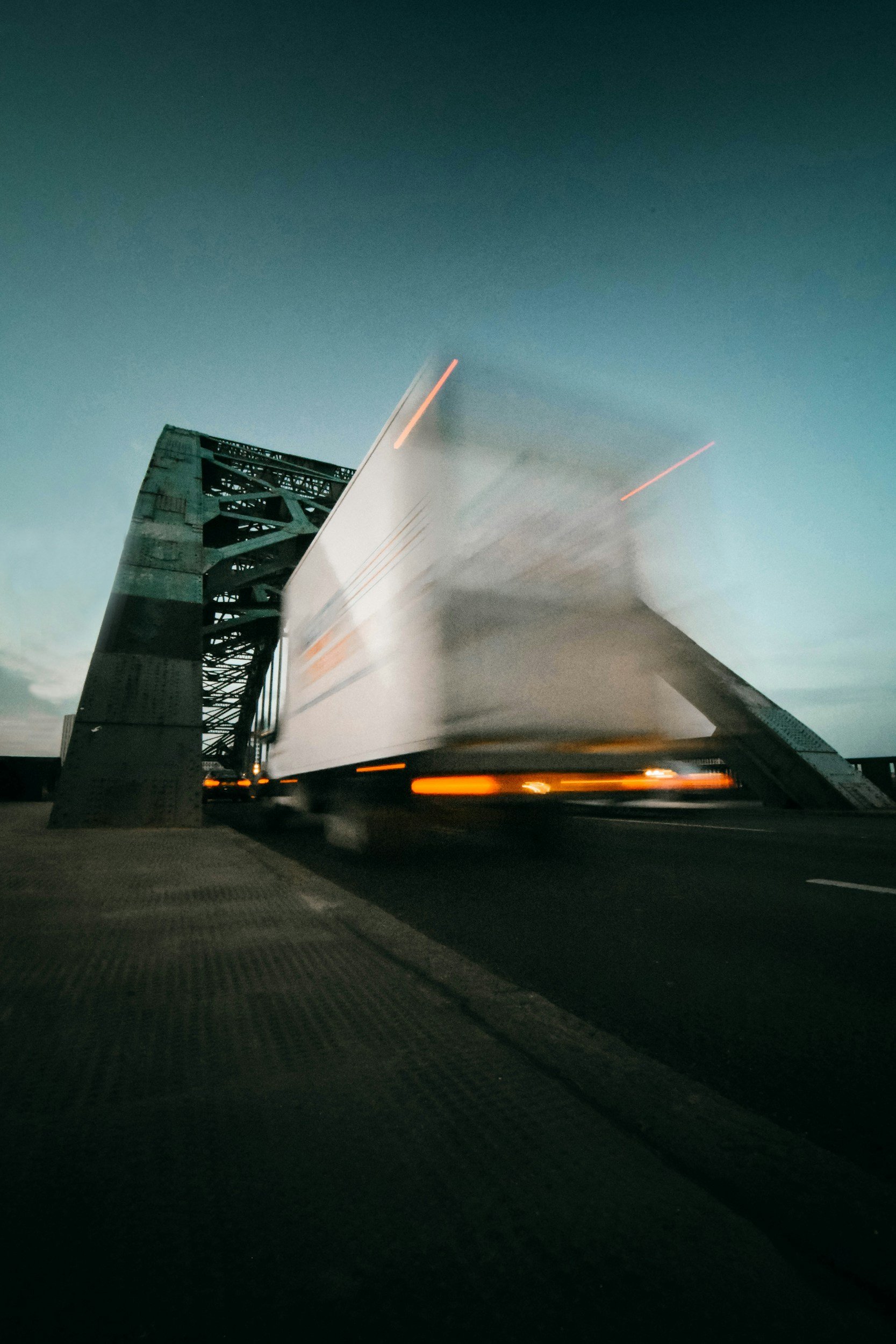 Blurred image of a truck crossing a steel bridge at dusk.