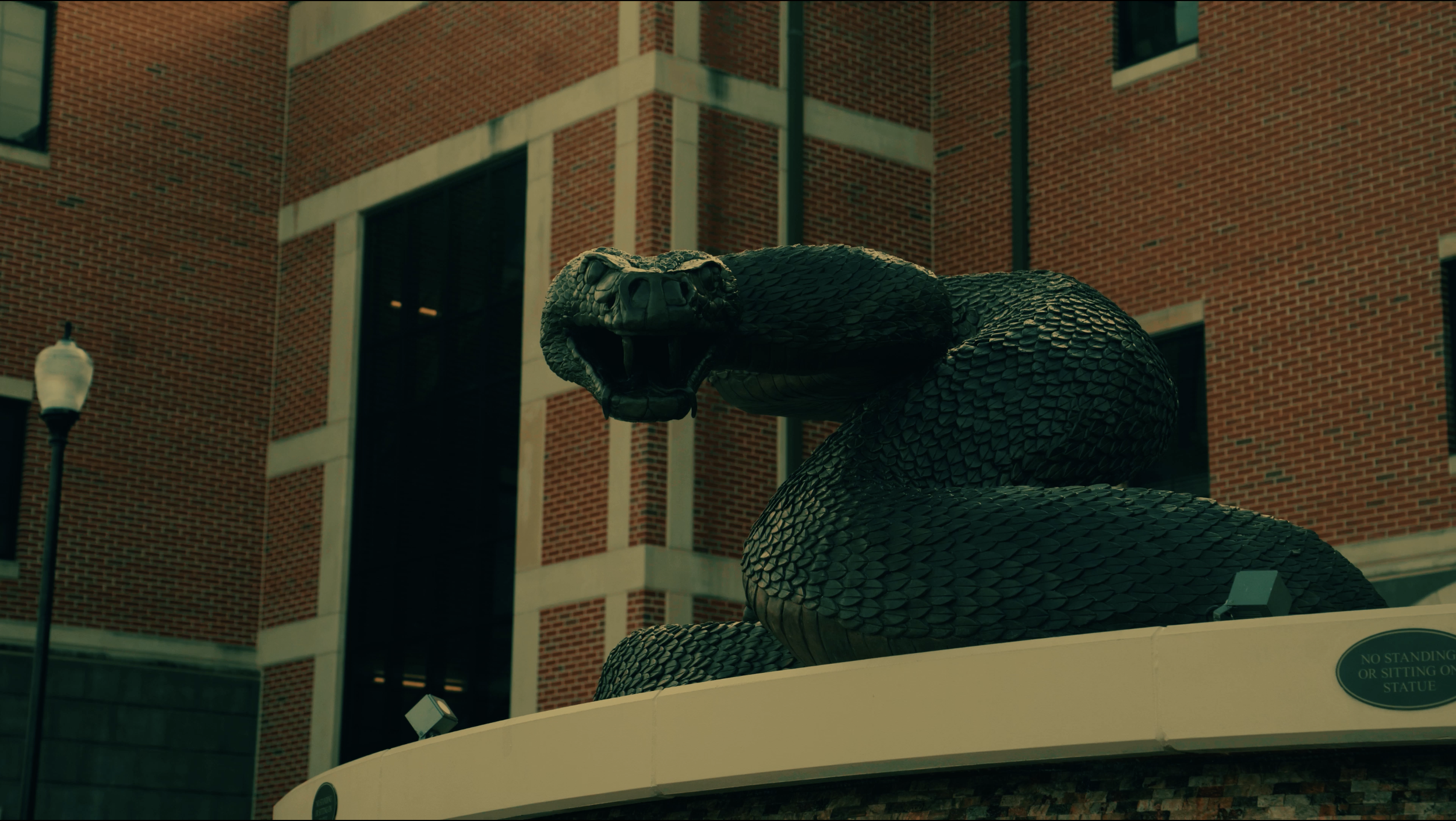 A large bronze snake sculpture with its mouth open, situated outdoors in front of a brick building with large windows.