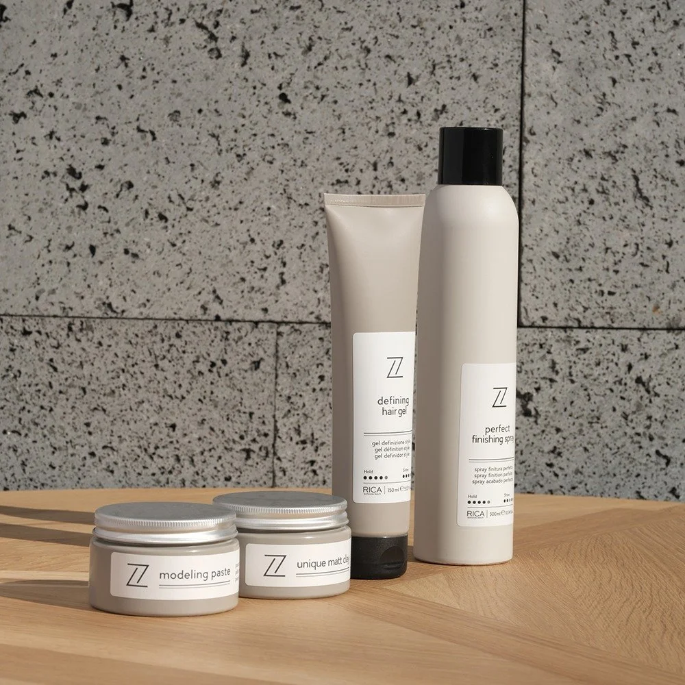 Produits de coiffure sur une table en bois devant un mur en pierre grise. Comprend un tube de gel coiffant, un spray de finition, et deux pots de pâte à modeler.