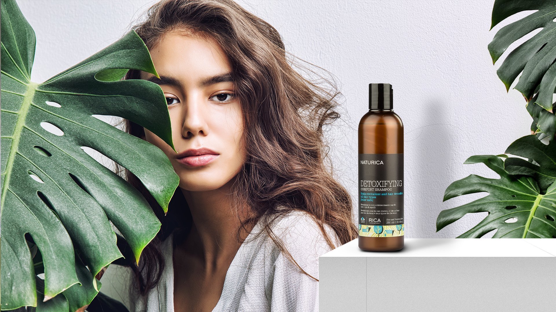 Une femme aux cheveux ondulés près de grandes feuilles de monstera dans une pièce neutre, avec une bouteille de shampoing naturel brun à l'arrière-plan sur un socle blanc.