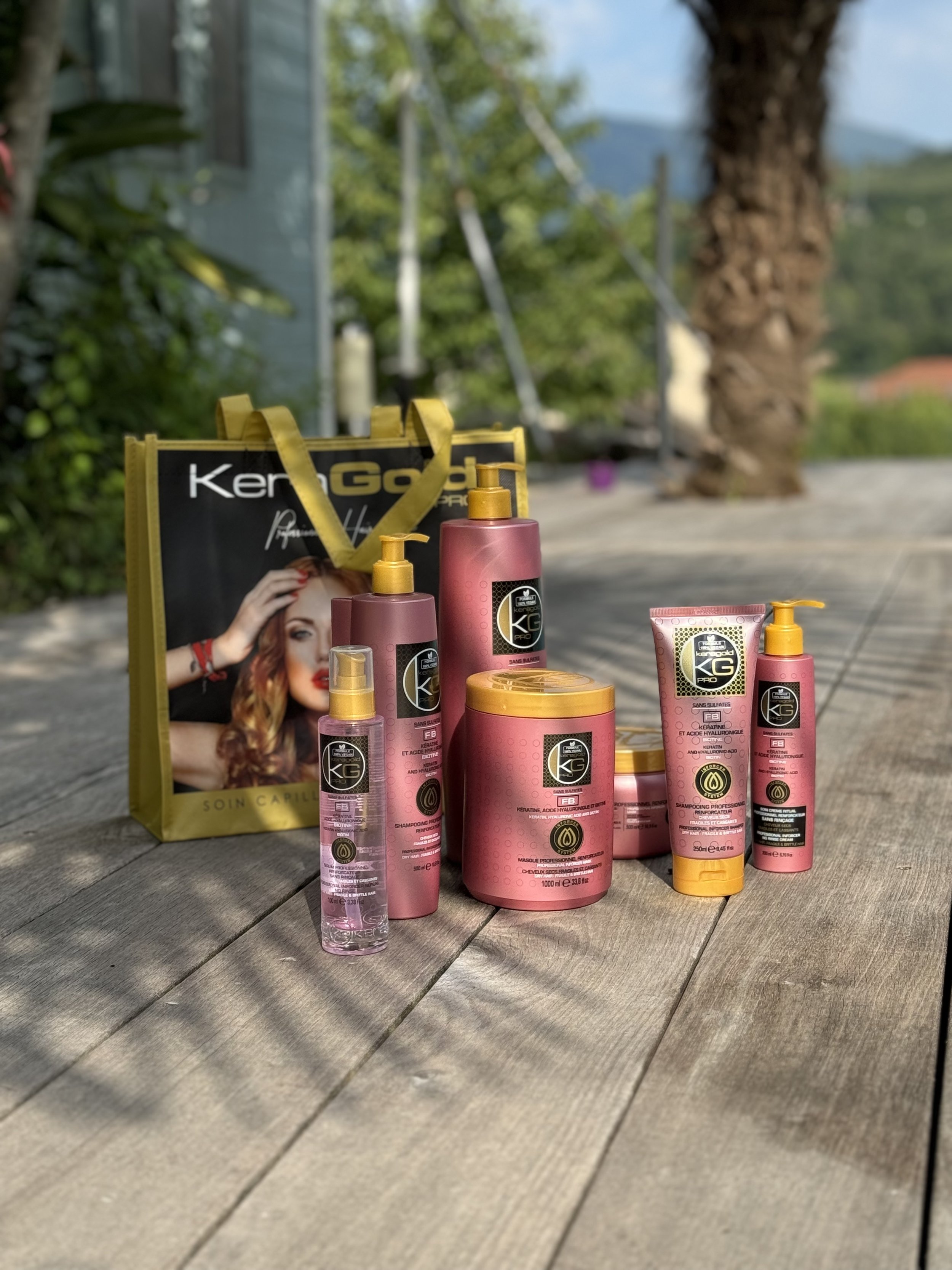 Une collection de produits capillaires en bouteilles roses et un sac de shopping noir et jaune avec une image d'une femme à cheveux roux, sur une surface en bois à l'extérieur, avec des arbres en arrière-plan.