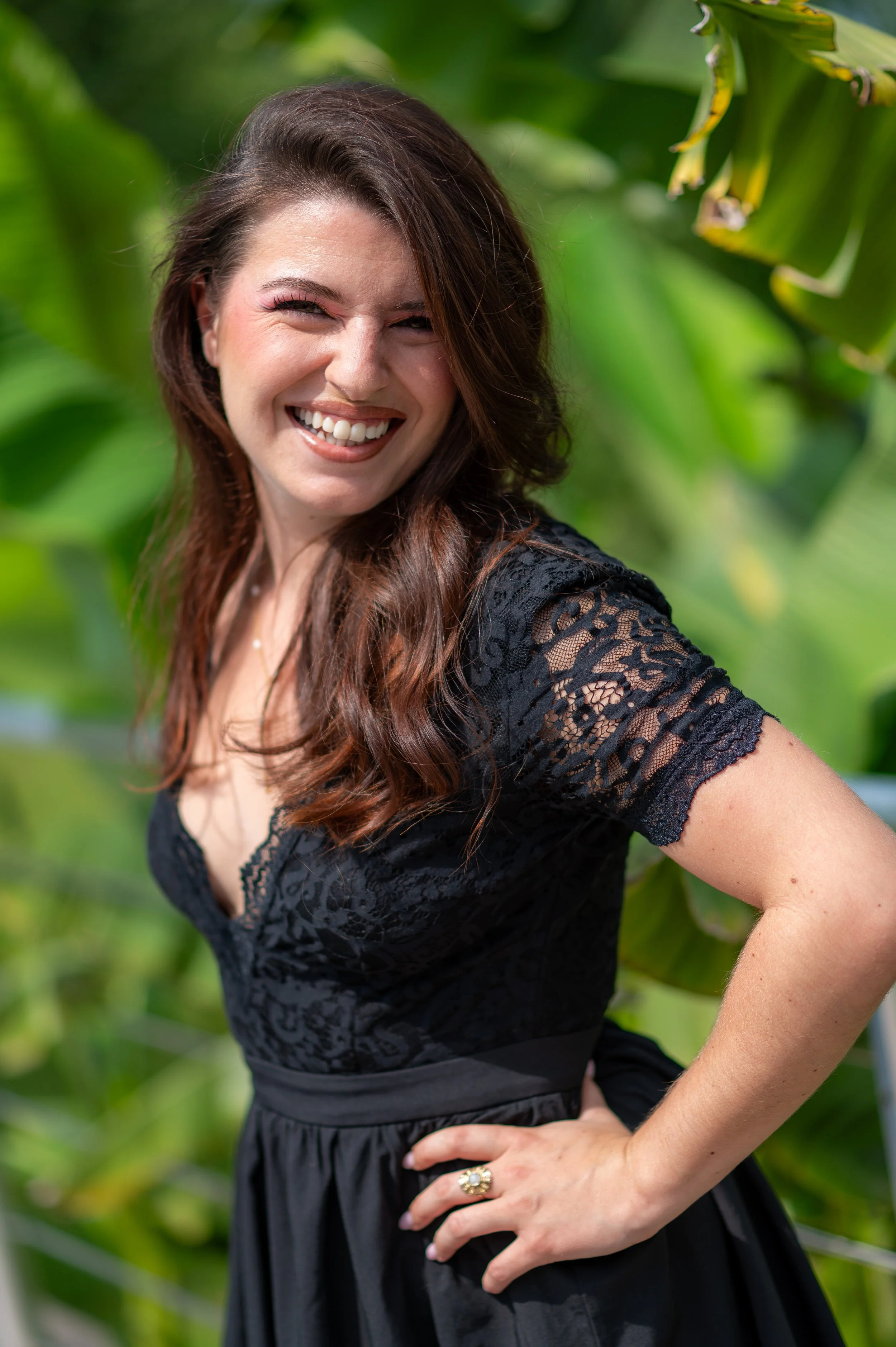 Une femme souriante portant une robe noire en dentelle, avec des feuilles vertes en arrière-plan.