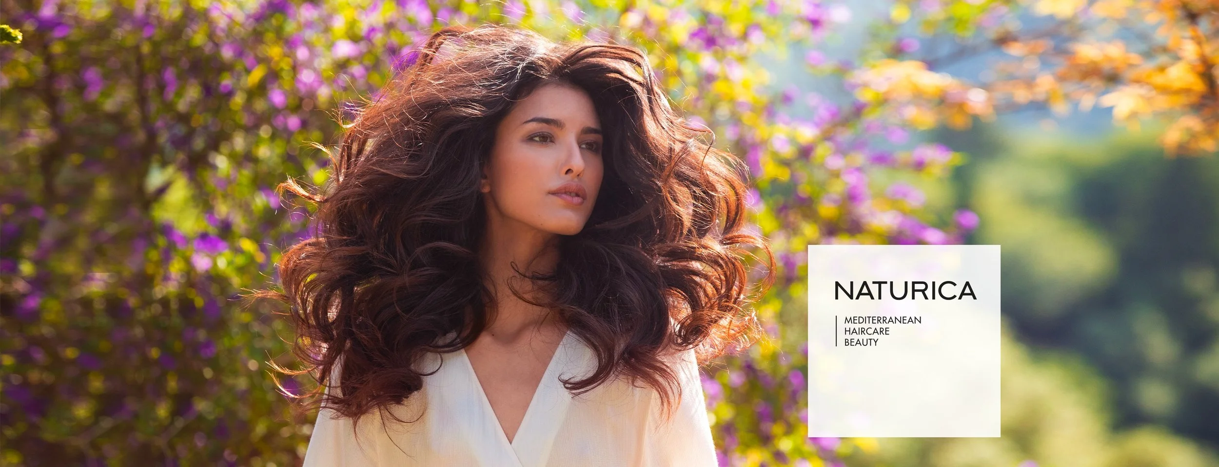 Femme aux cheveux longs et bouclés devant un fond de nature avec des fleurs violettes et du feuillage vert. Texte sur l'image : 'NATURICA, MÉDITERRANÉEN, COIFFURE, BEAUTÉ'.