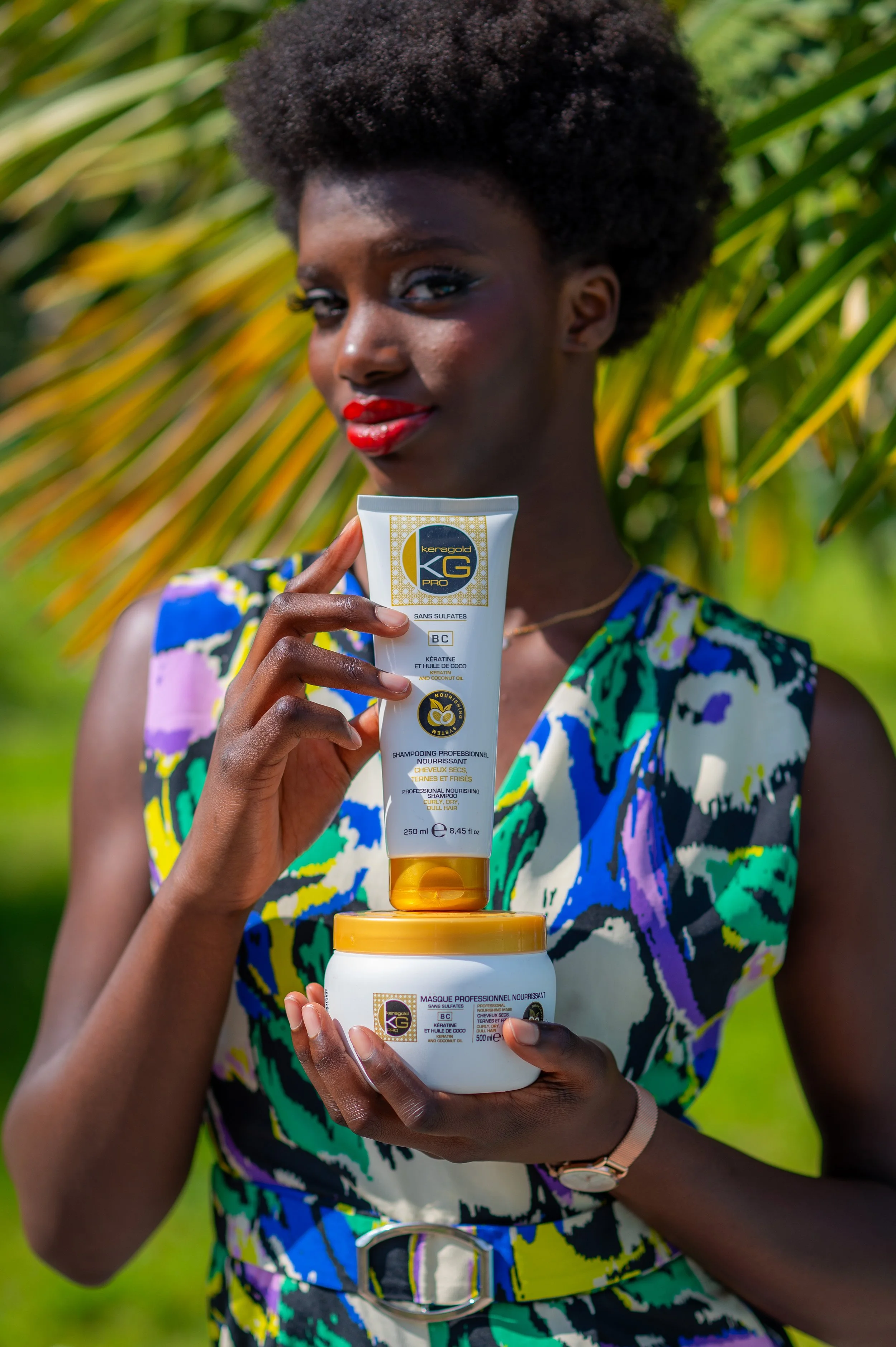 Une femme noire avec des cheveux bouclés courts, portant une robe colorée, tient deux produits capillaires. Elle se trouve à l’extérieur, parmi des feuilles vertes, et regarde l'appareil photo avec un sourire.