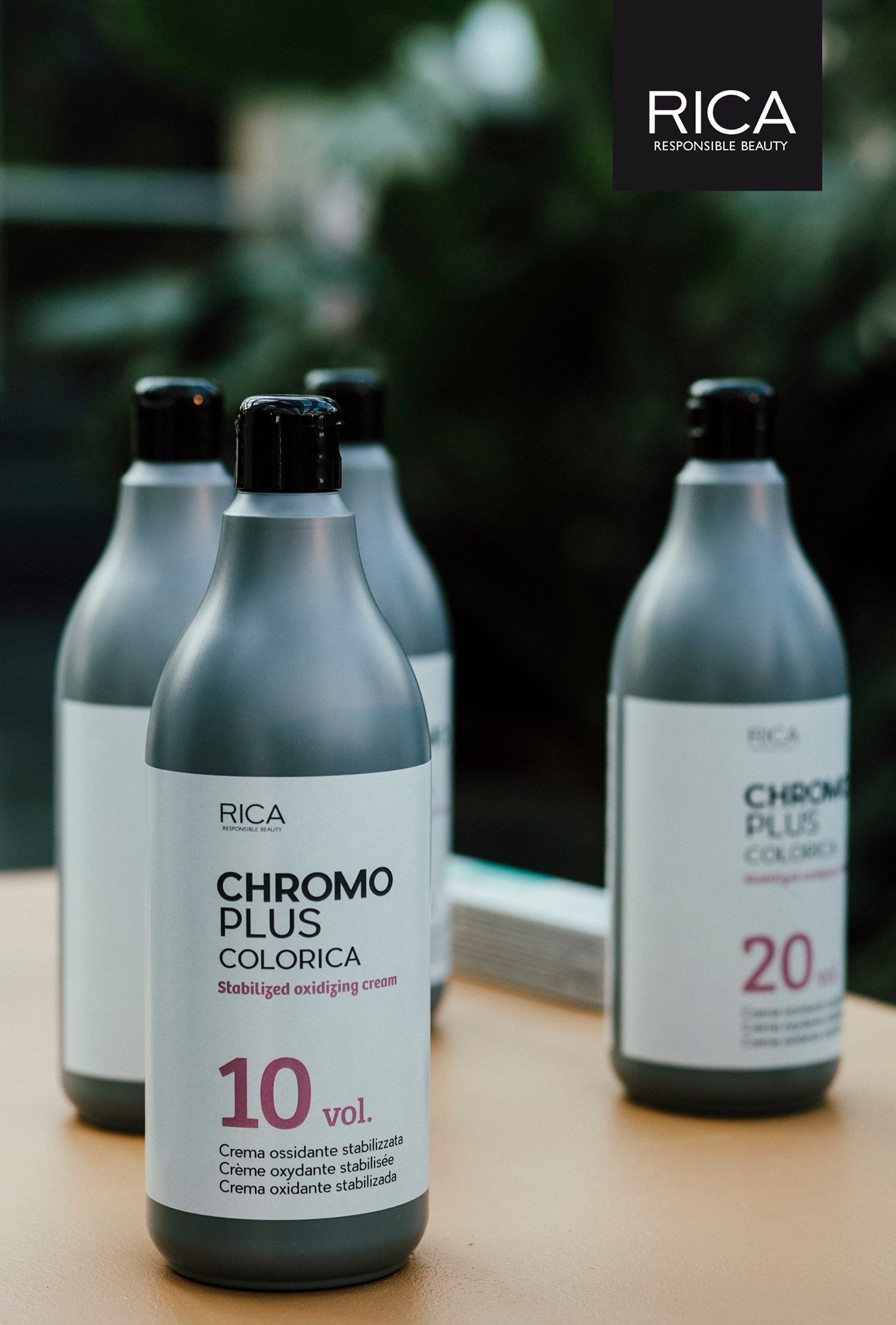 Trois bouteilles de crème colorante pour cheveux sur une table en bois, avec des étiquettes indiquant des volumes 10 et 20, la marque RICA et le produit Chromo Plus Colorica. Fond flou d'un espace naturel ou botanique.