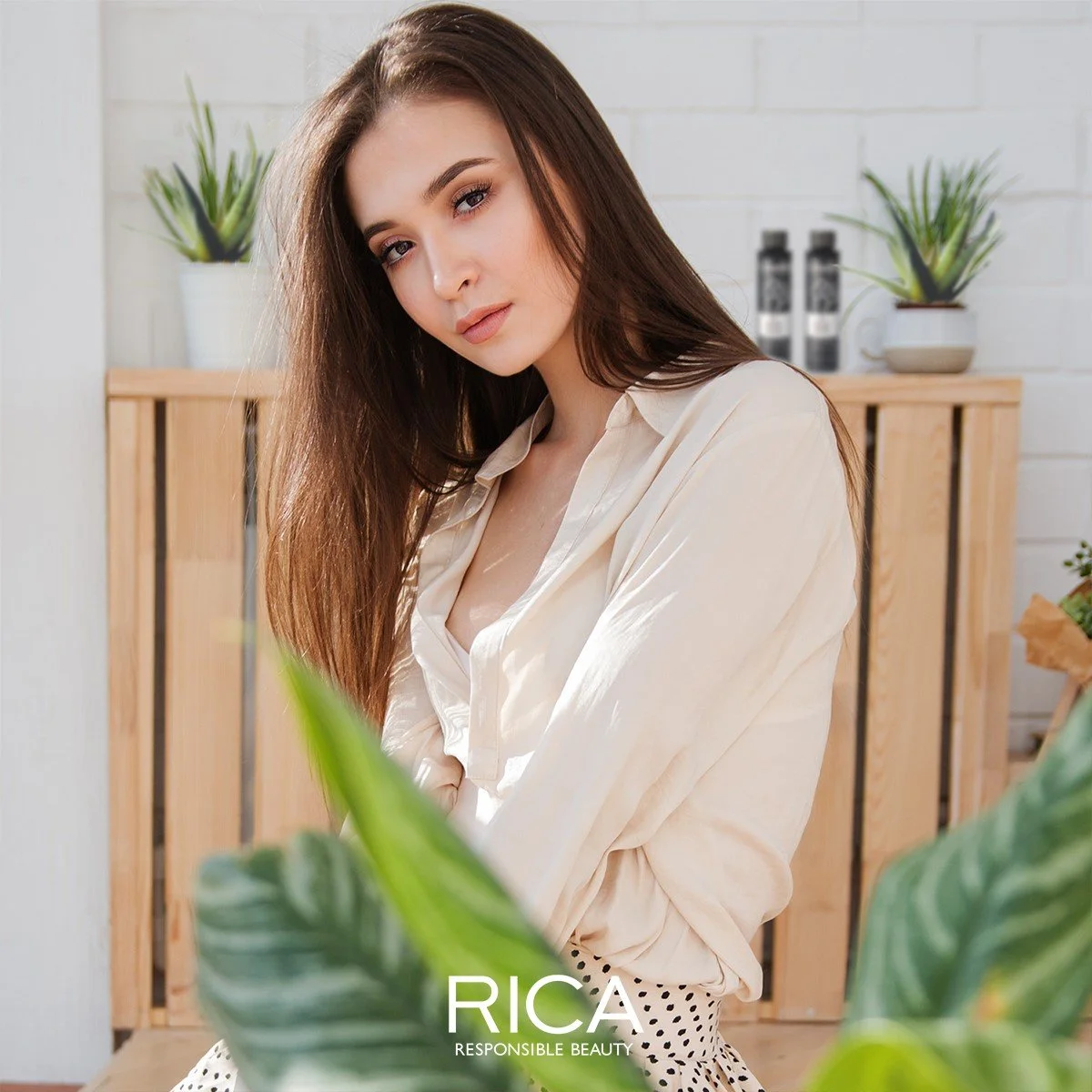 Une jeune femme aux longs cheveux bruns porte une chemise beige clair dans un intérieur lumineux avec des plantes vertes, en arrière-plan une étagère en bois et des potsatalogues.