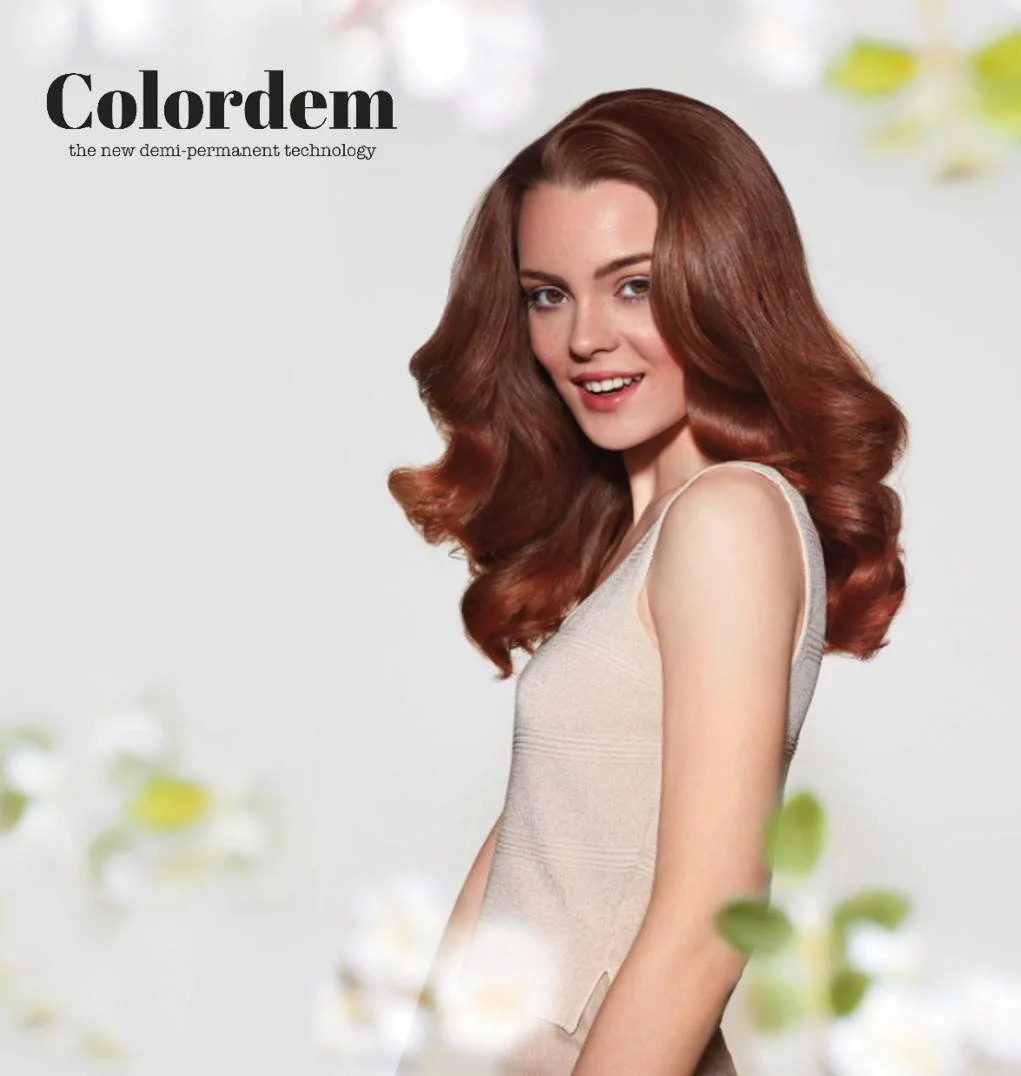 Une femme avec de longs cheveux bouclés de couleur châtain rouge, portant une robe beige, souriant et regardant vers l'avant, avec un fond clair et quelques feuilles floues en premier plan.