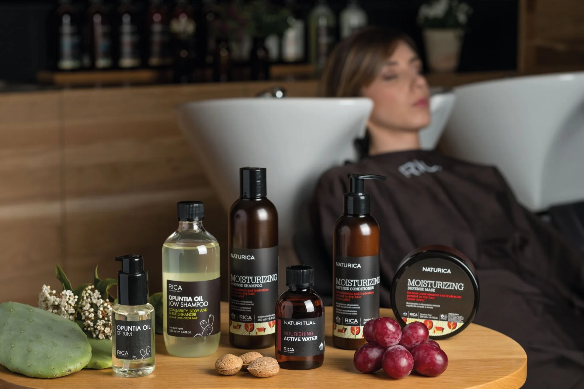 Produits de soin capillaire et des ingrédients naturels sur une table en bois, avec une femme se faisant laver ou traiter les cheveux en arrière-plan dans un salon de coiffure.