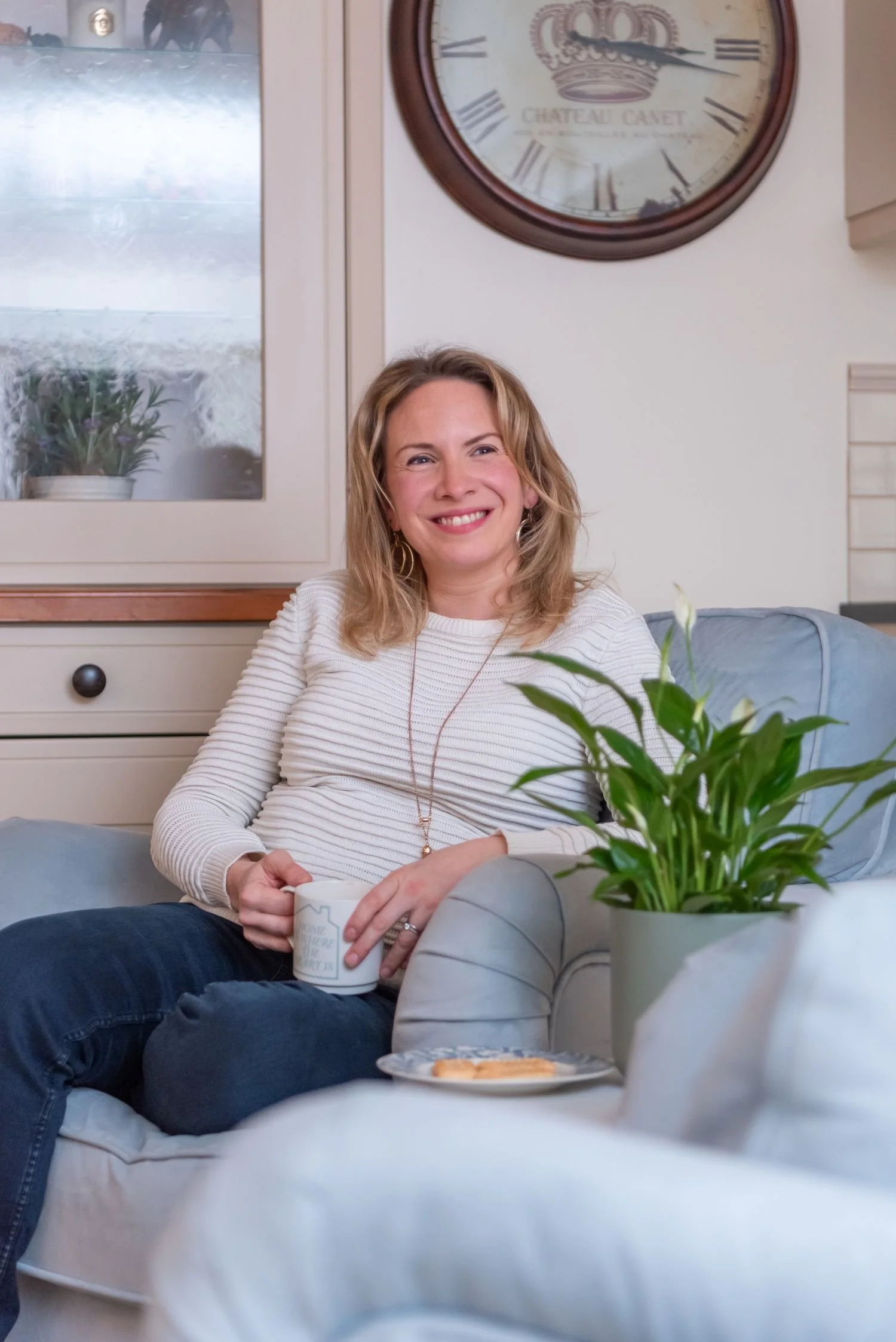 A woman with blonde hair, smiling, sitting on a comfy chair in a cozy living room, holding a mug, with a plant and a plate of cookies next to her, and a large wall clock behind her.