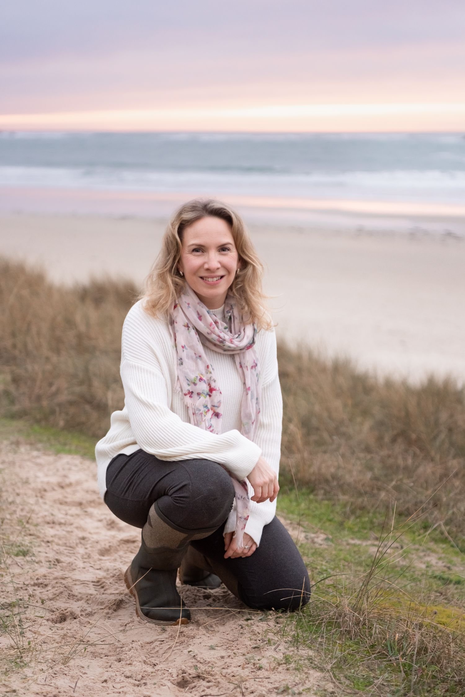 A woman with blonde hair smiling and squatting on a beach at sunset, wearing a white sweater, a floral scarf, black pants, and boots, with the ocean and a colorful sky in the background.