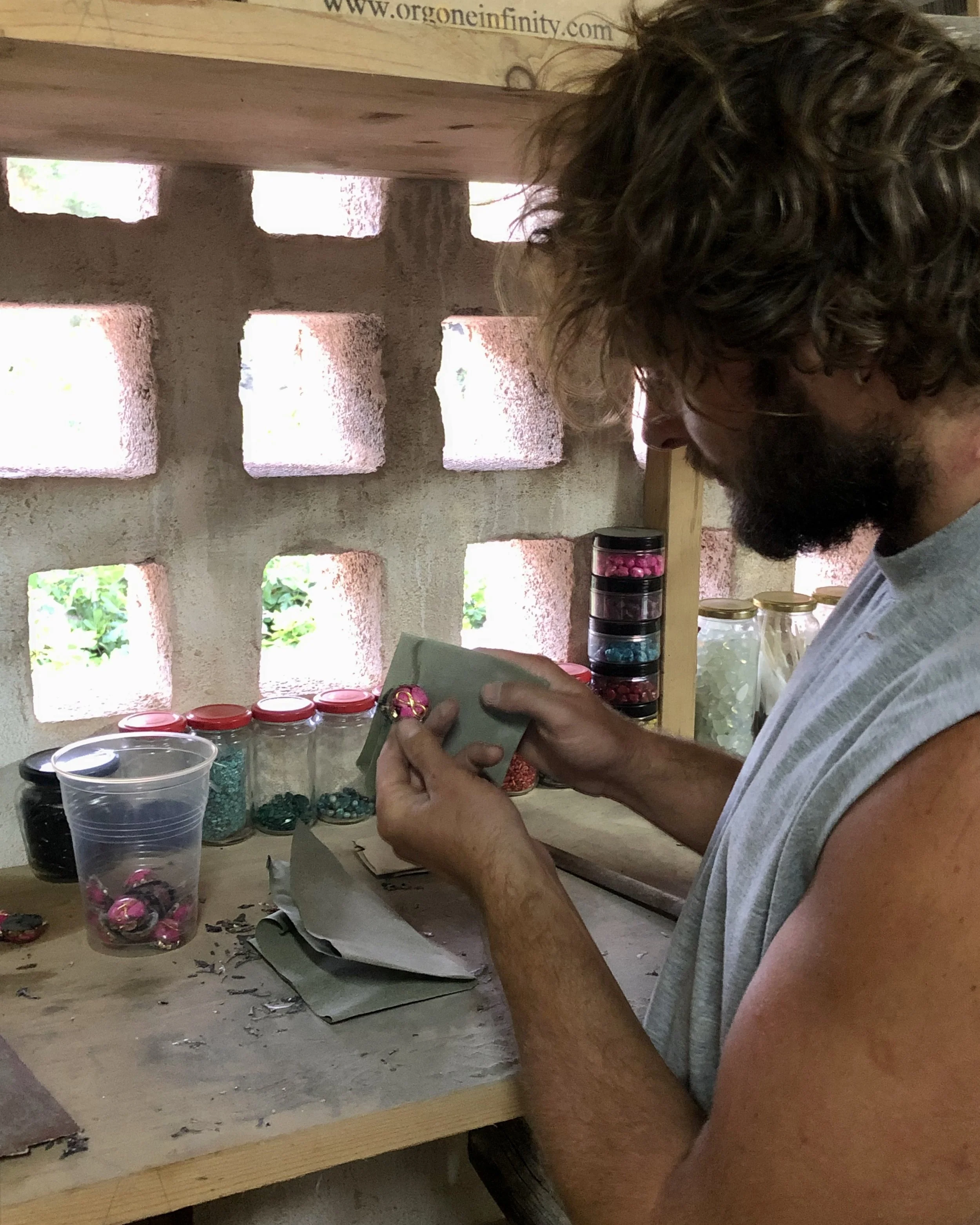 Man with curly hair and beard sitting at a wooden workbench, working with small colorful objects, jars of beads, and craft supplies in a workshop with concrete block walls.