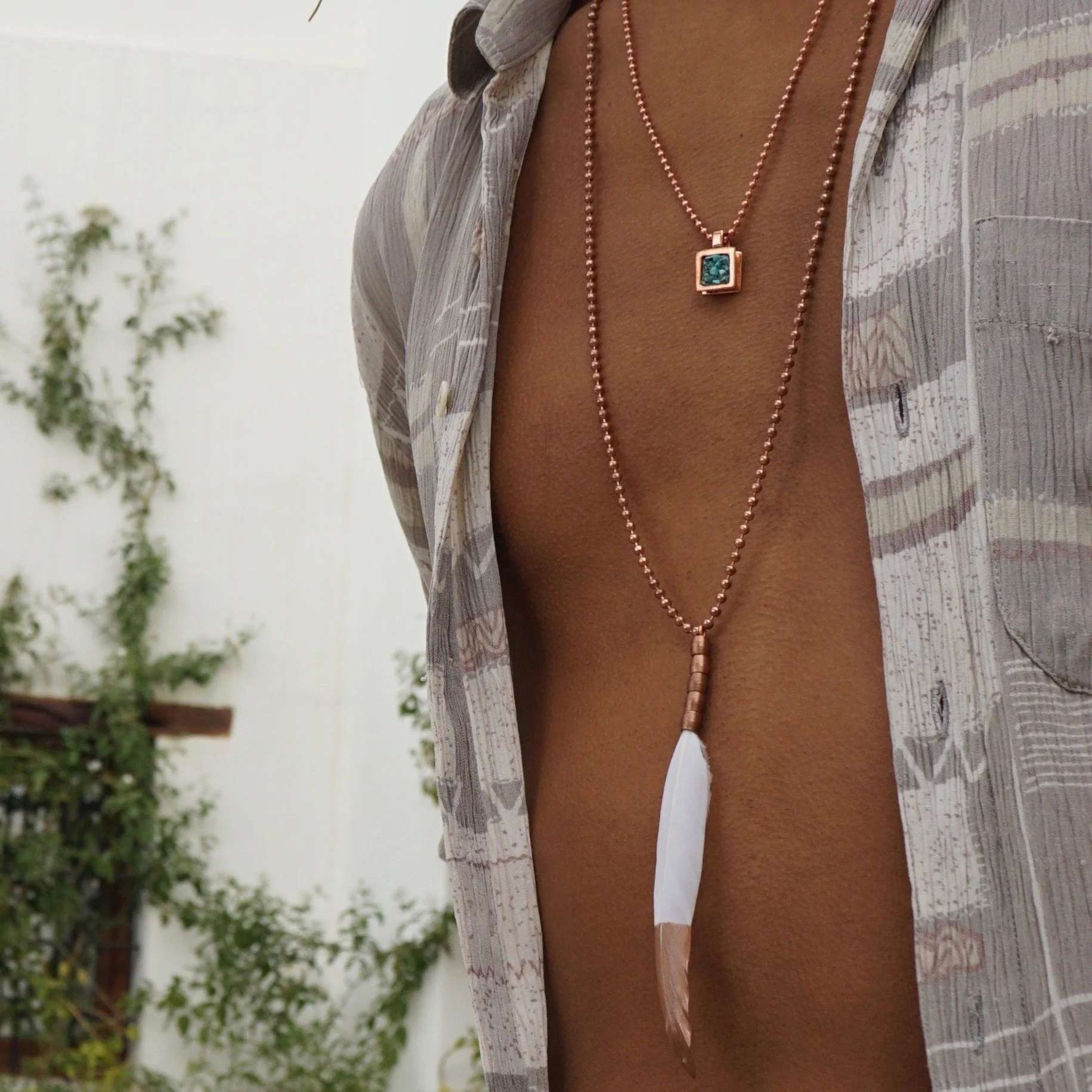 Close-up of a person's chest and neck, wearing layered necklaces with beads, a square pendant, and a feather, open shirt, and exposed skin.