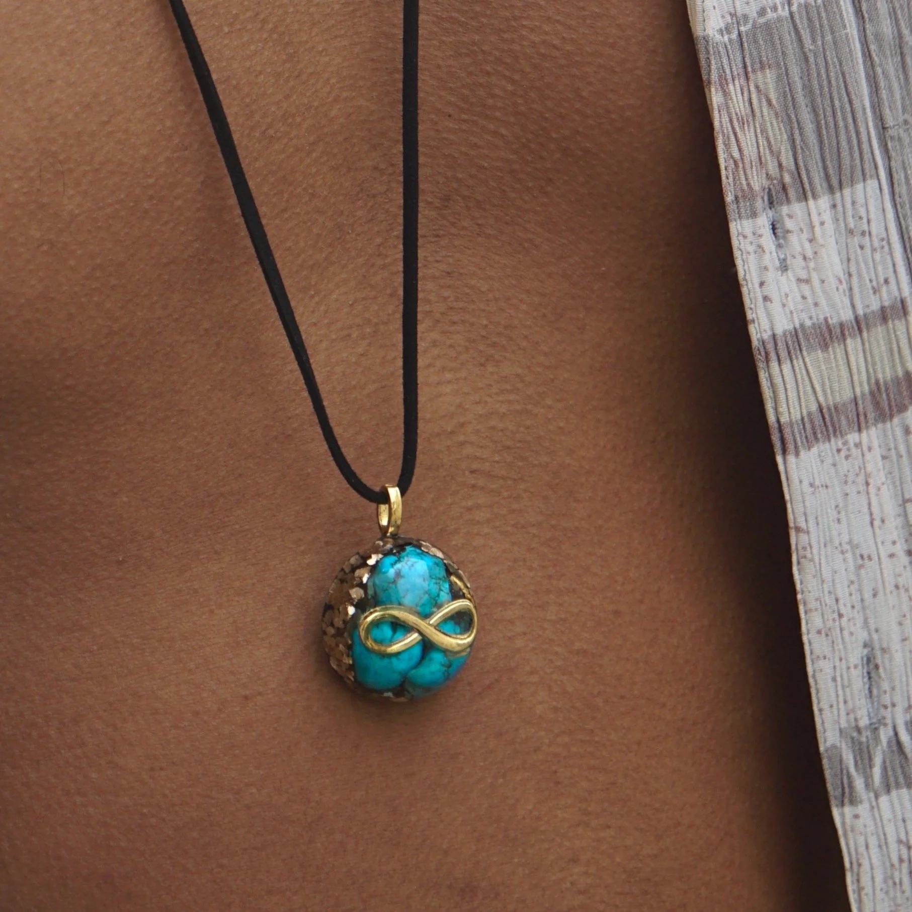 Necklace with a turquoise stone pendant and gold infinity symbol on a black cord, worn by a person with brown skin and wearing a light-colored, patterned shirt.