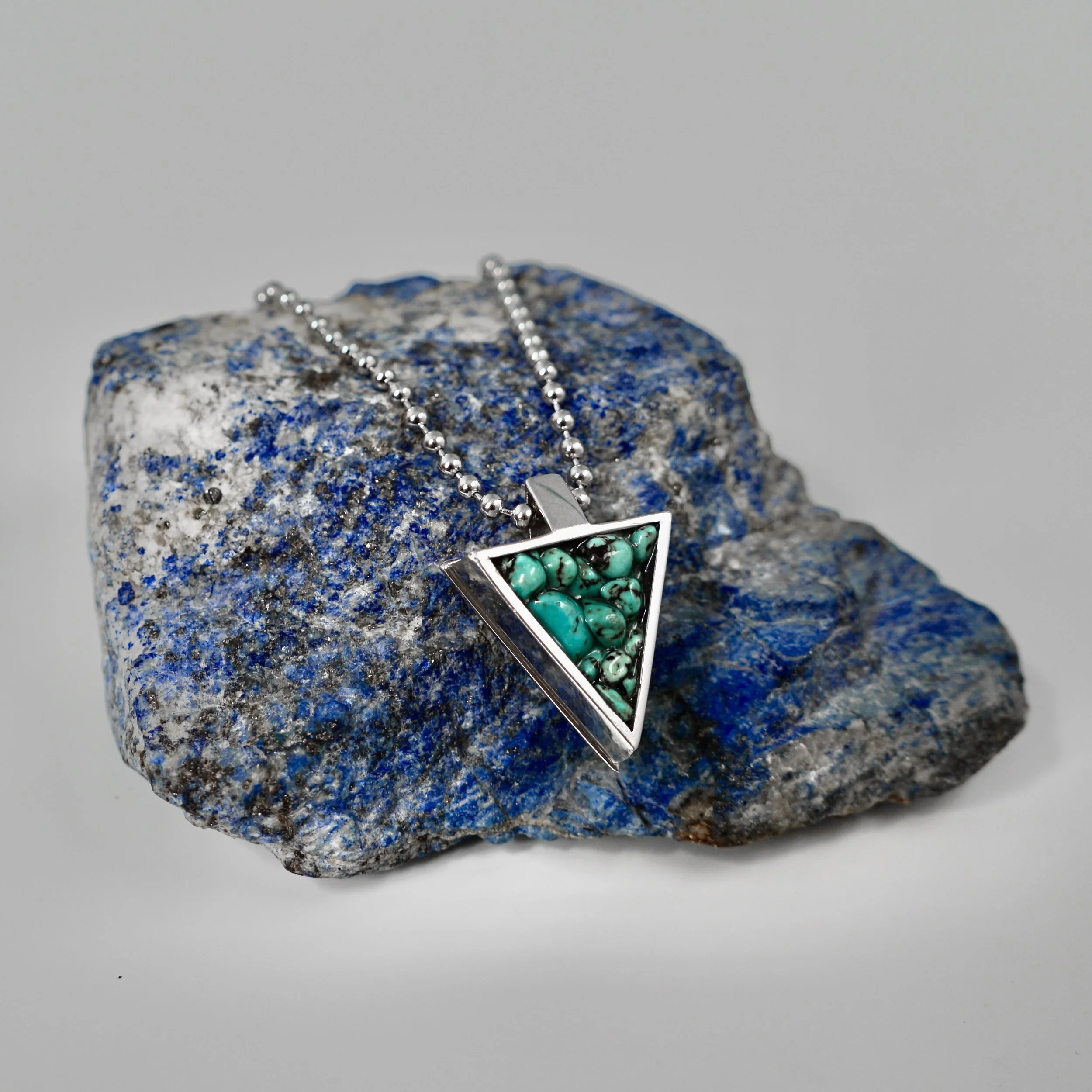 Silver necklace with a triangular pendant filled with turquoise stones, resting on a blue and gray stone.
