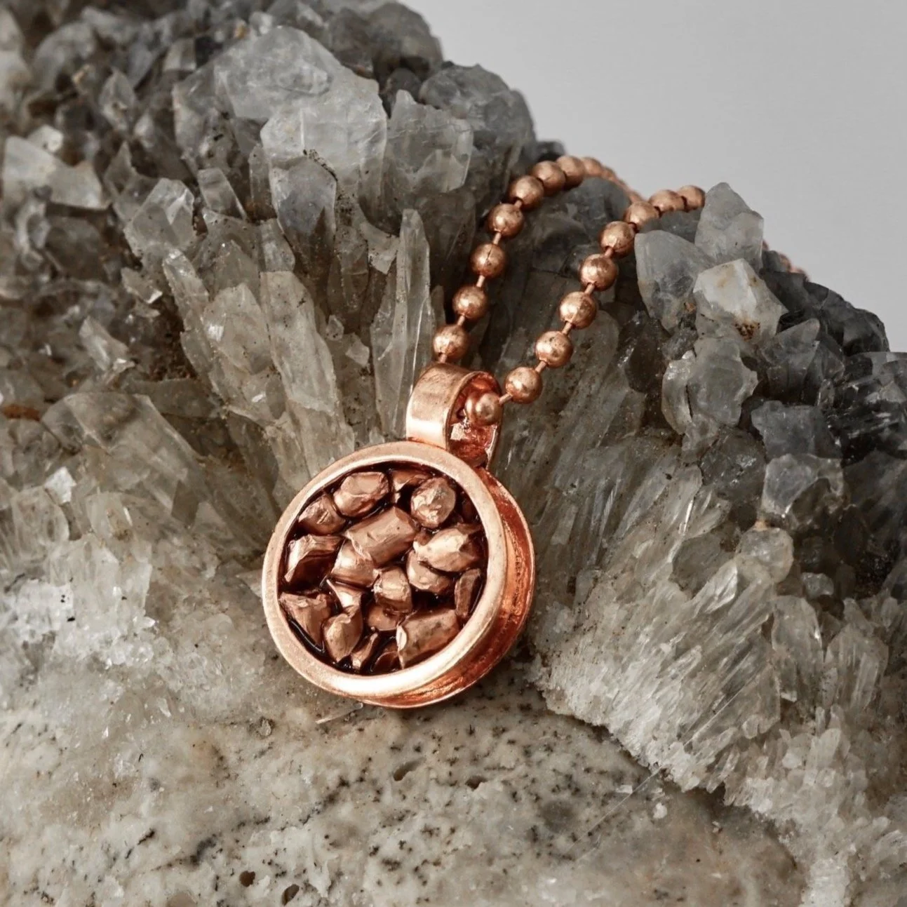 A rose gold pendant necklace with small beads hanging on a chain, placed on a cluster of clear quartz crystals.
