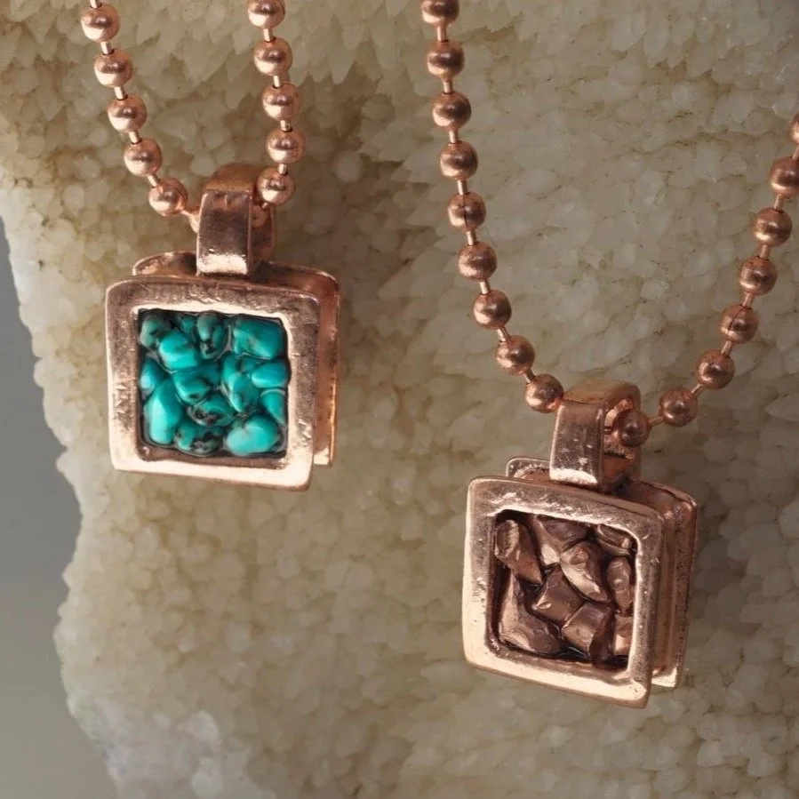 Two gold square pendants on gold ball chains, one with turquoise stones and the other with brown stones, hanging against a textured cream background.