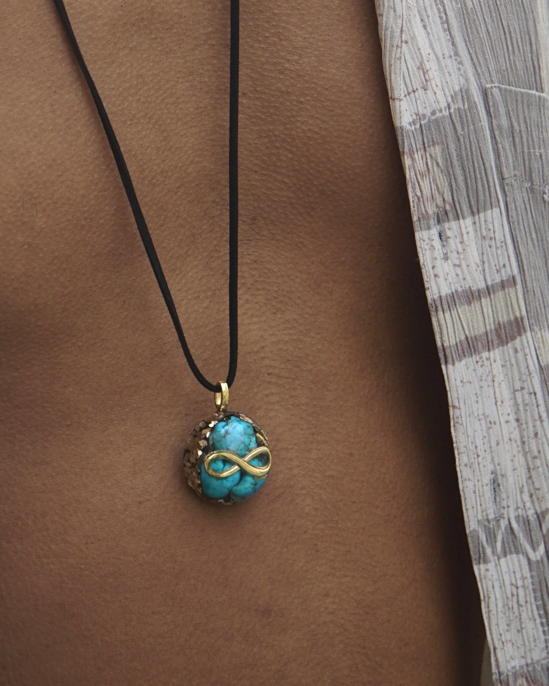 Close-up of a person's chest with a turquoise gemstone pendant necklace featuring a gold infinity symbol on a black cord, partially covered by a striped shirt.