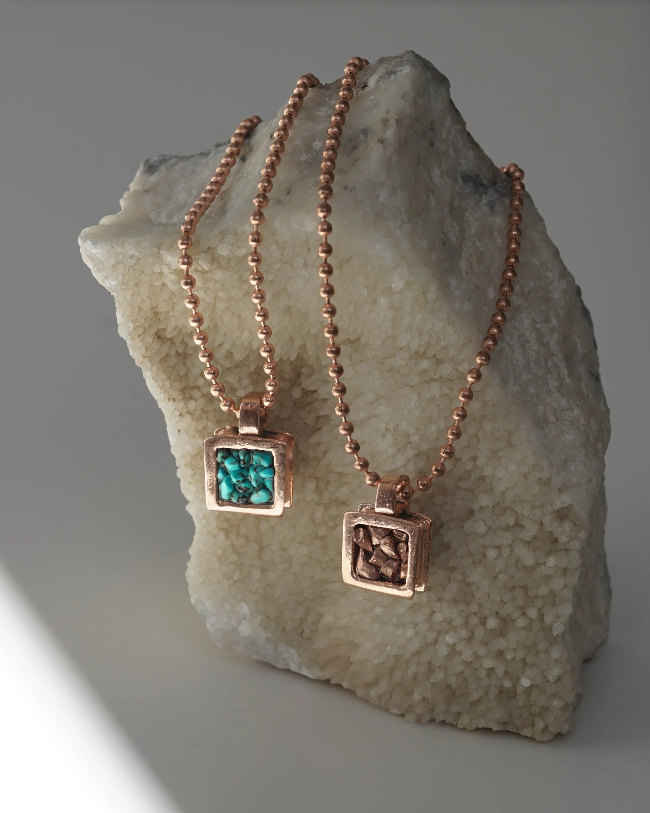 Two necklaces with square pendants, one with turquoise stones and the other with a carved brown stone, displayed on a textured white stone background.