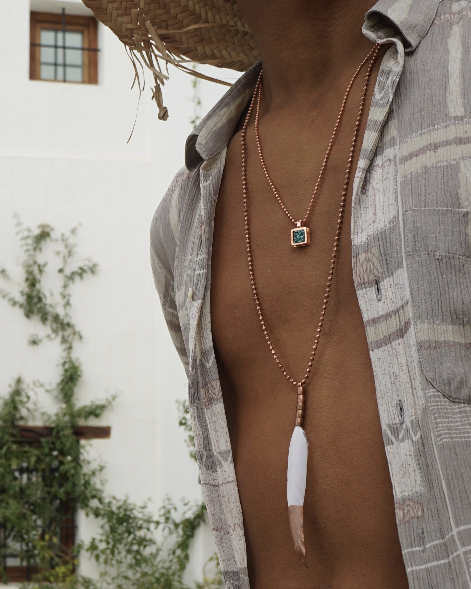 Person wearing necklaces, open shirt, straw hat, and standing outdoors with house and plants in background.