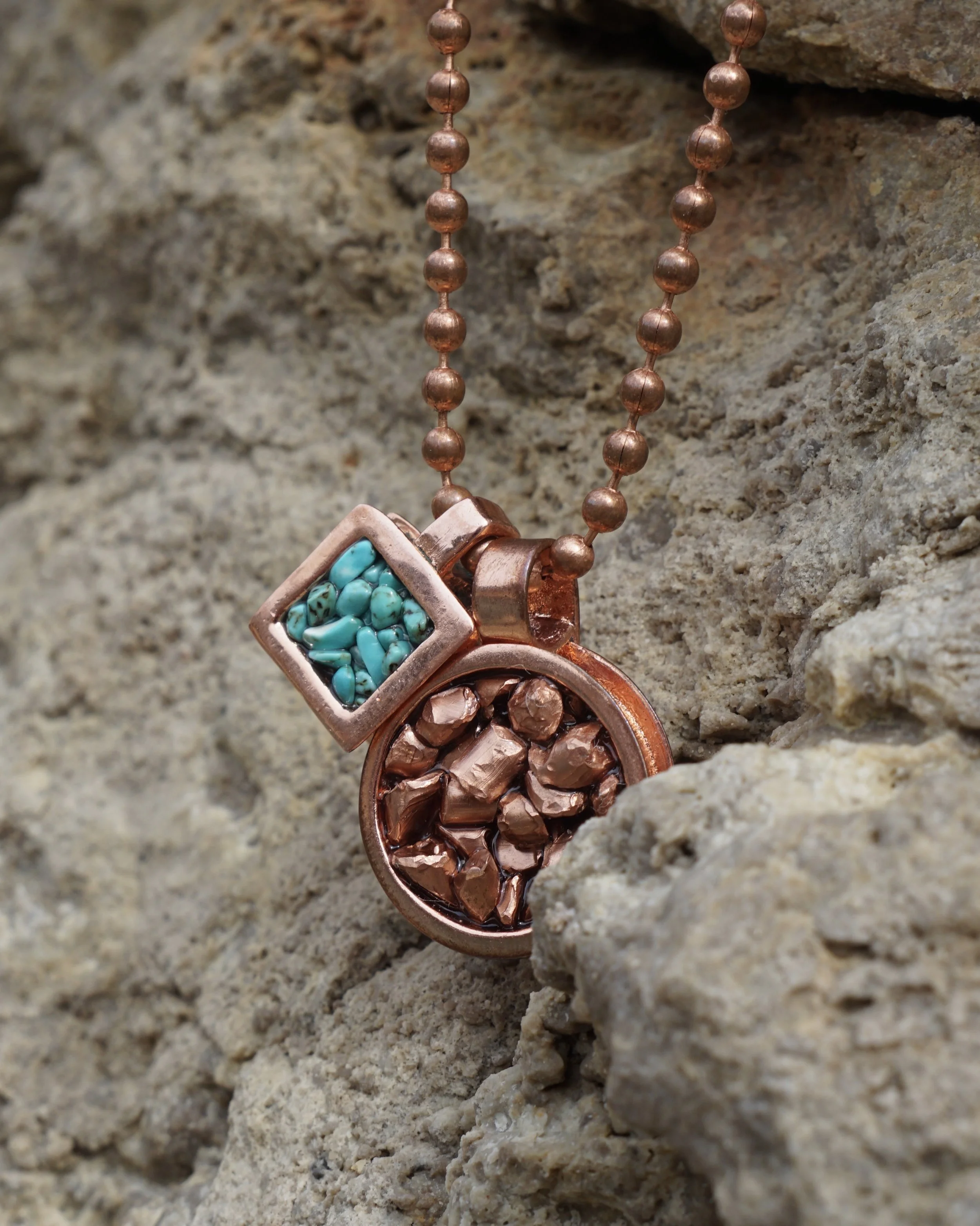 A copper necklace with two pendants, one square with turquoise stones and one round with copper chunks, hanging against a rough stone background.