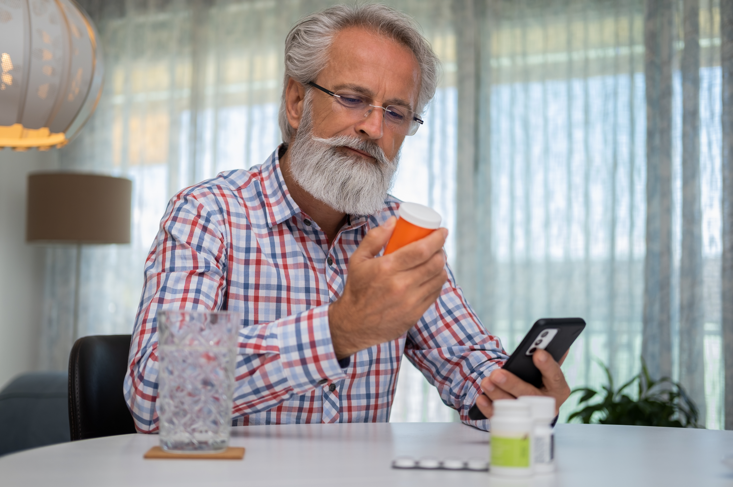 An elderly man with gray hair and beard, wearing glasses and a checkered shirt, looking at a prescription bottle while holding a smartphone. There are other pill bottles and a glass of water on the table.