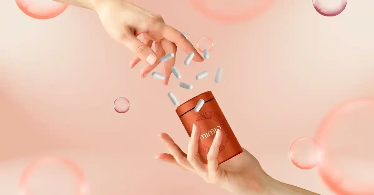 Hands holding a small orange container dispensing white capsules against a pink background with floating bubbles